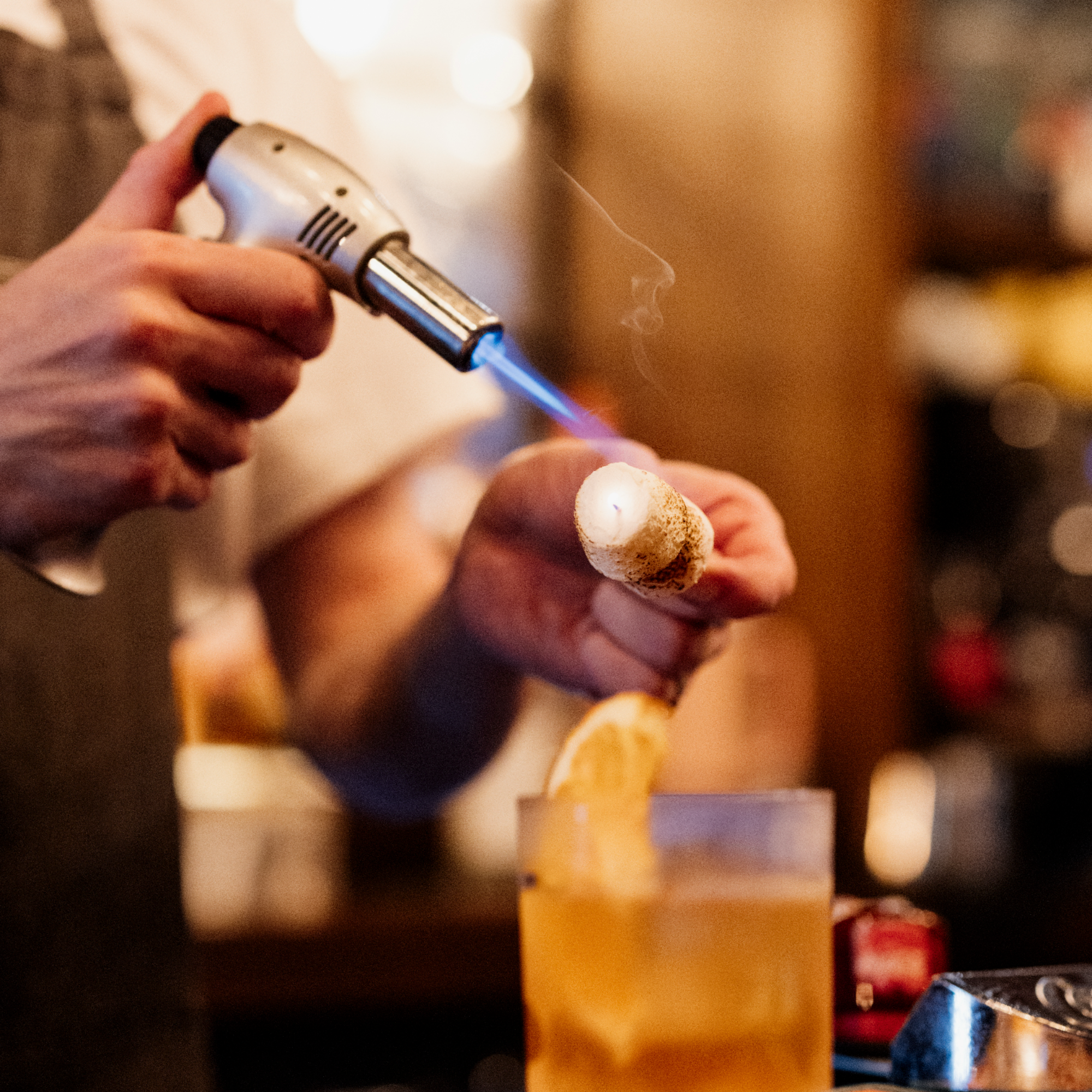 A person holding a lighter with a blue flame, igniting a marshmallow on a skewer over a fire.