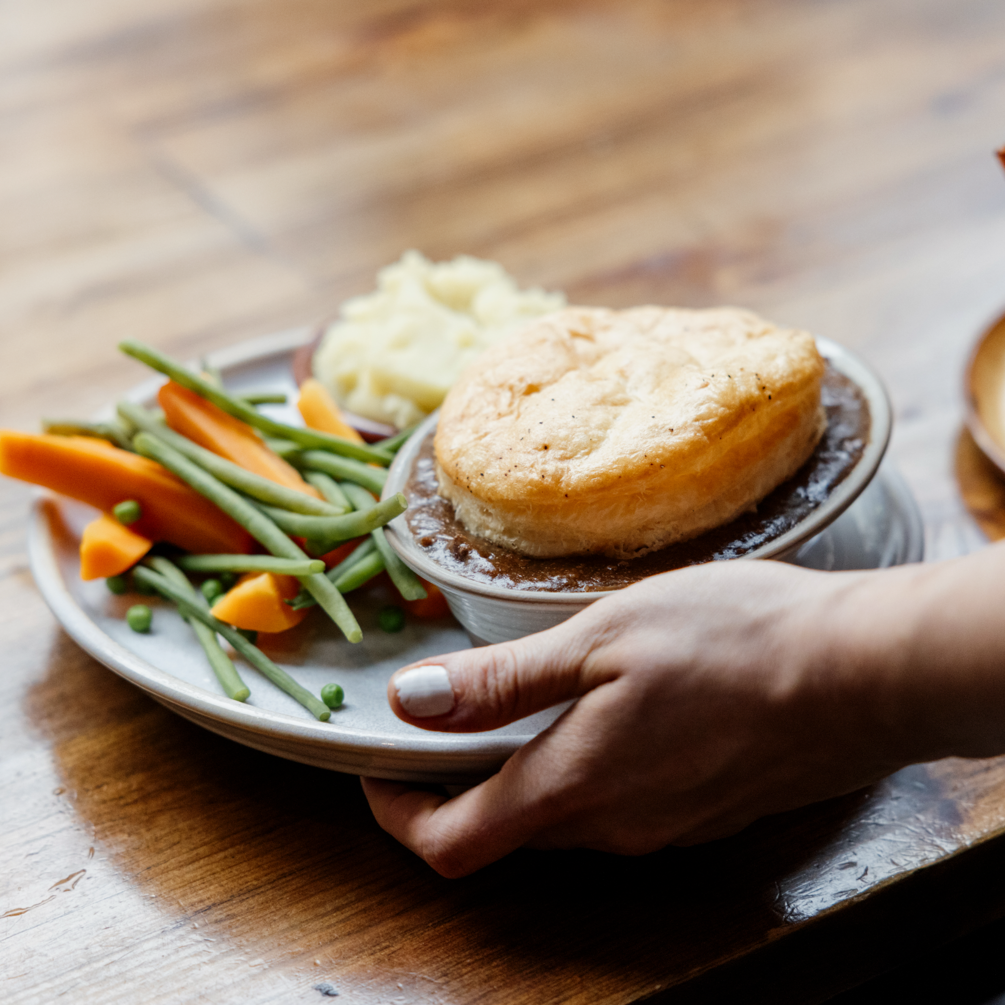 A hand holding a plate of steak pie, with a plate of mixed vegetables nearby, on a wooden table.
