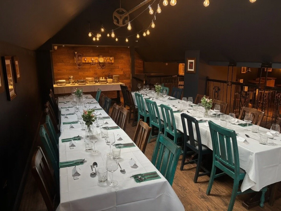 A decorated dining area in a restaurant with tables set with white tablecloths, glassware, silverware, and floral centerpieces, illuminated by hanging string lights. Private hire and mezzanine area.