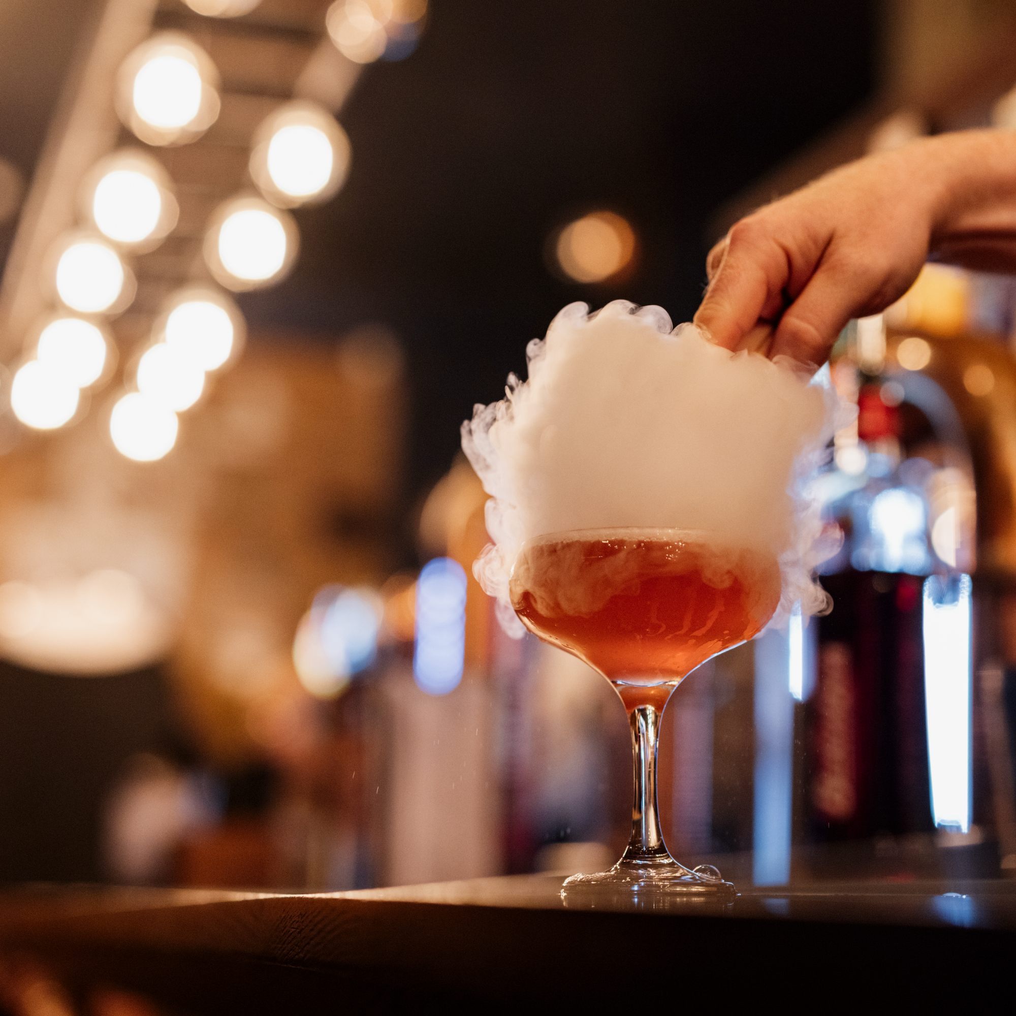 A close-up of a cocktail glass filled with an orange-colored beverage, topped with smoke, with a hand reaching for the glass in a blurry, brightly-lit background.