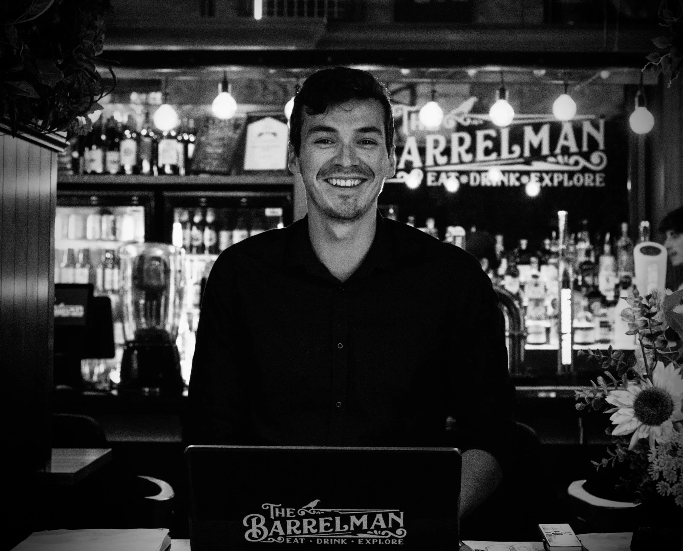 A smiling man standing in front of a bar with a sign that reads "The Barrelman Eat Drink Explore," with bottles of alcohol on shelves behind him and decorated with hanging lights.