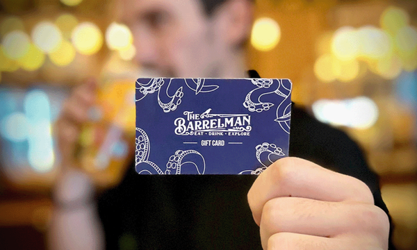Person holding a gift card from The Barrelman  with blurred background of lights and a drink.