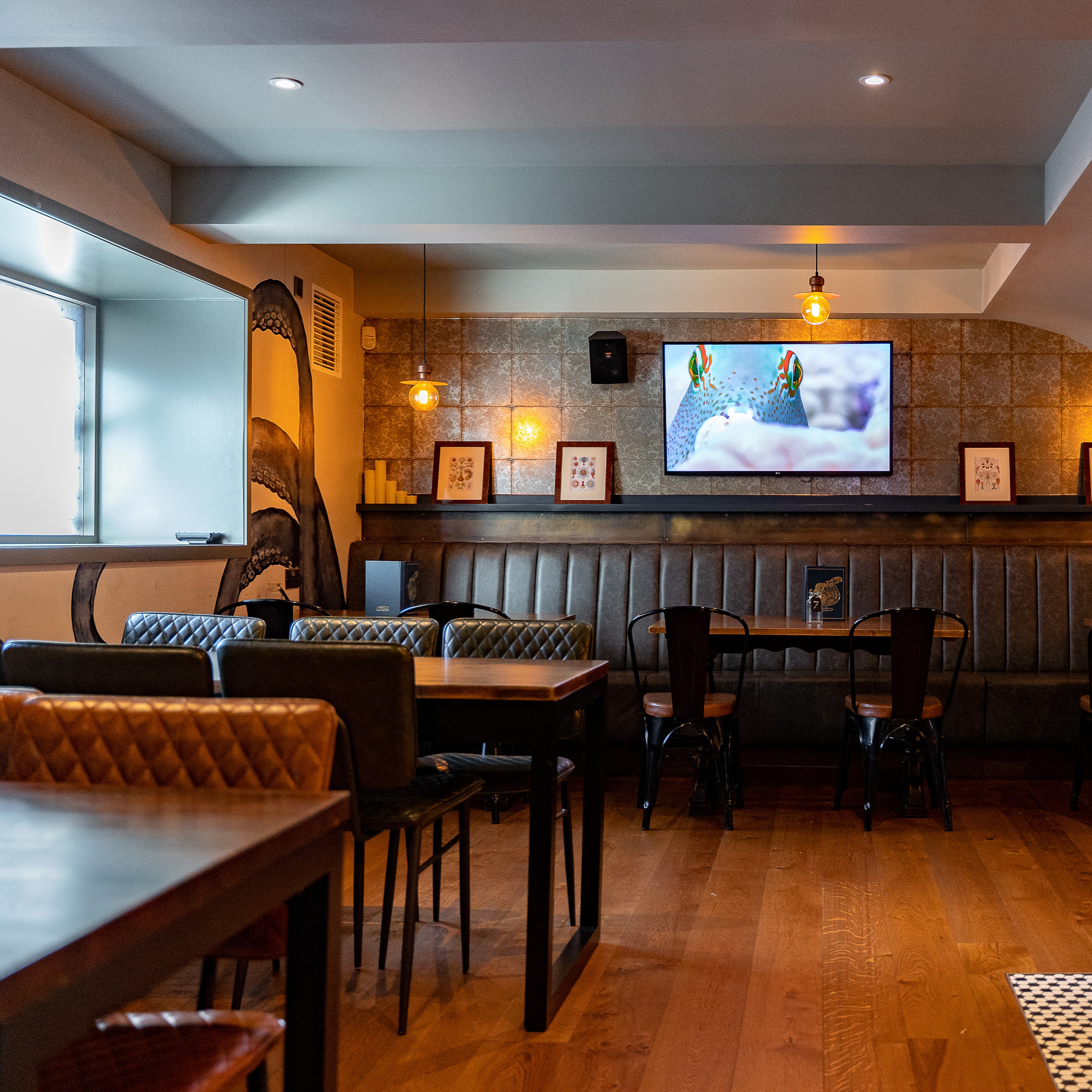 Interior of a restaurant or cafe with wooden tables and chairs, a TV displaying an image of a fish, and warm lighting from hanging bulbs.