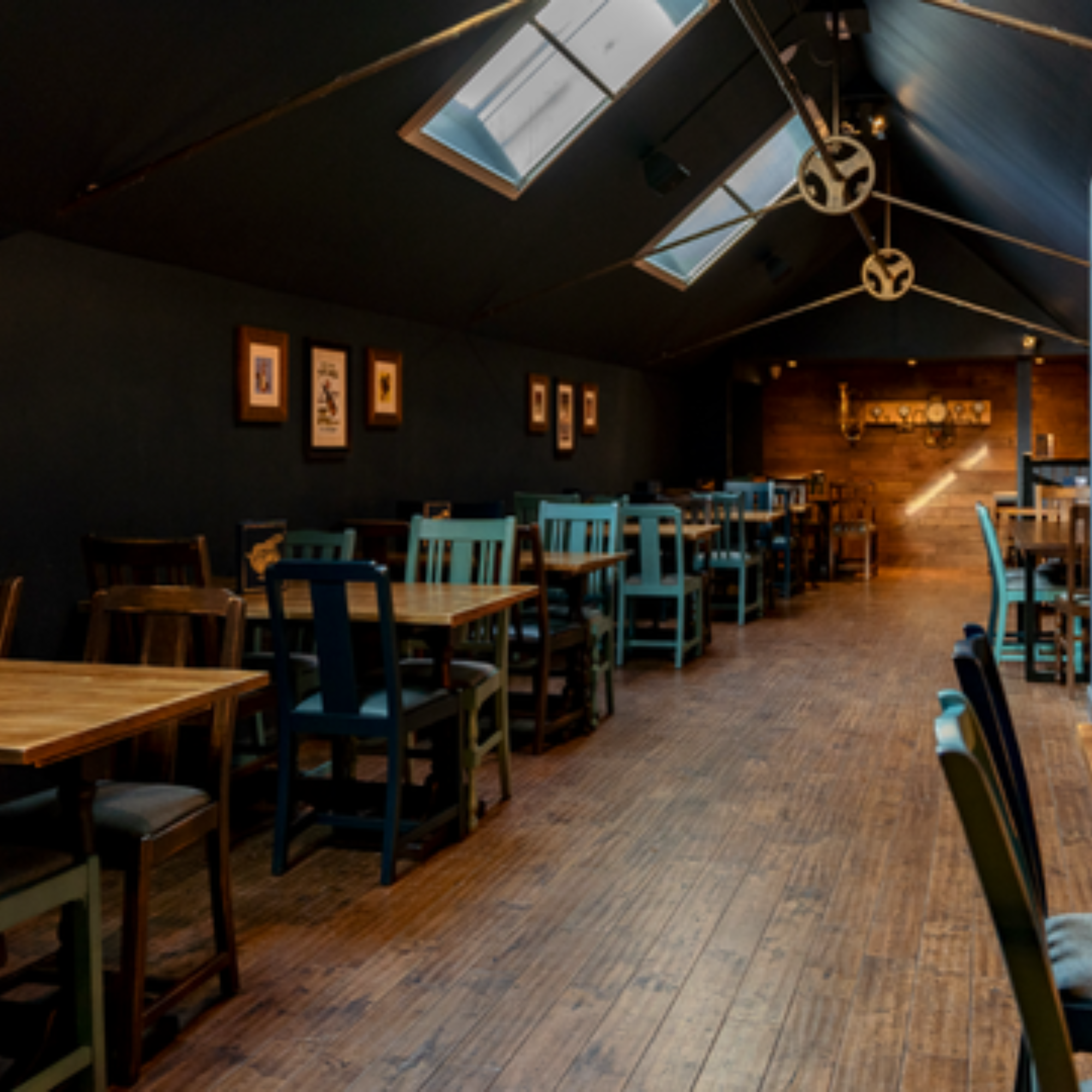 Interior of a restaurant on mezzanine with wooden floors, blue and brown chairs, and a dark wall decorated with framed pictures. Skylights provide natural light.