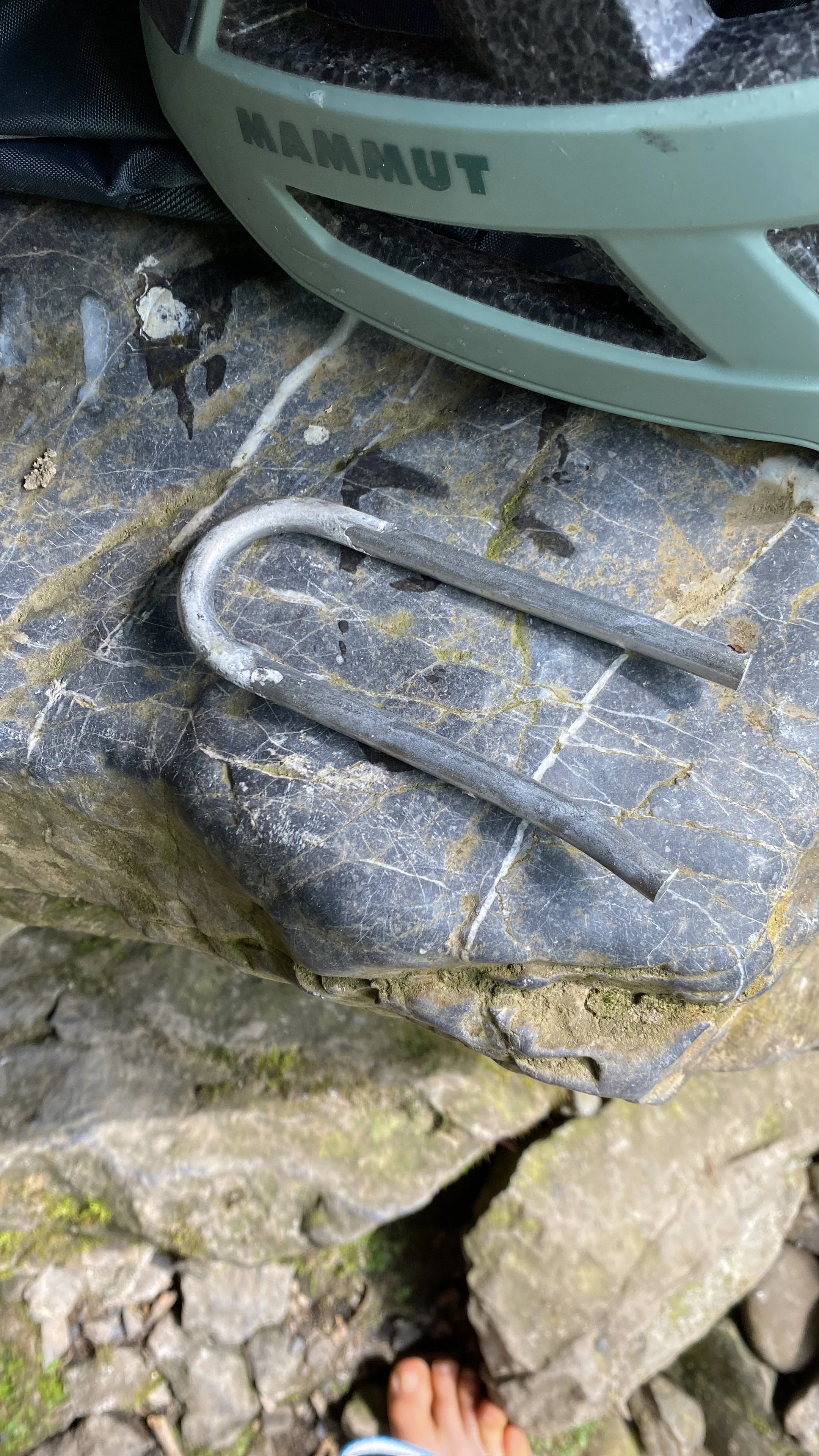 A close-up of a rock surface with a metal hook and a U-lock. A green helmet with the word 'MAMMUT' is partially visible at the top of the image.