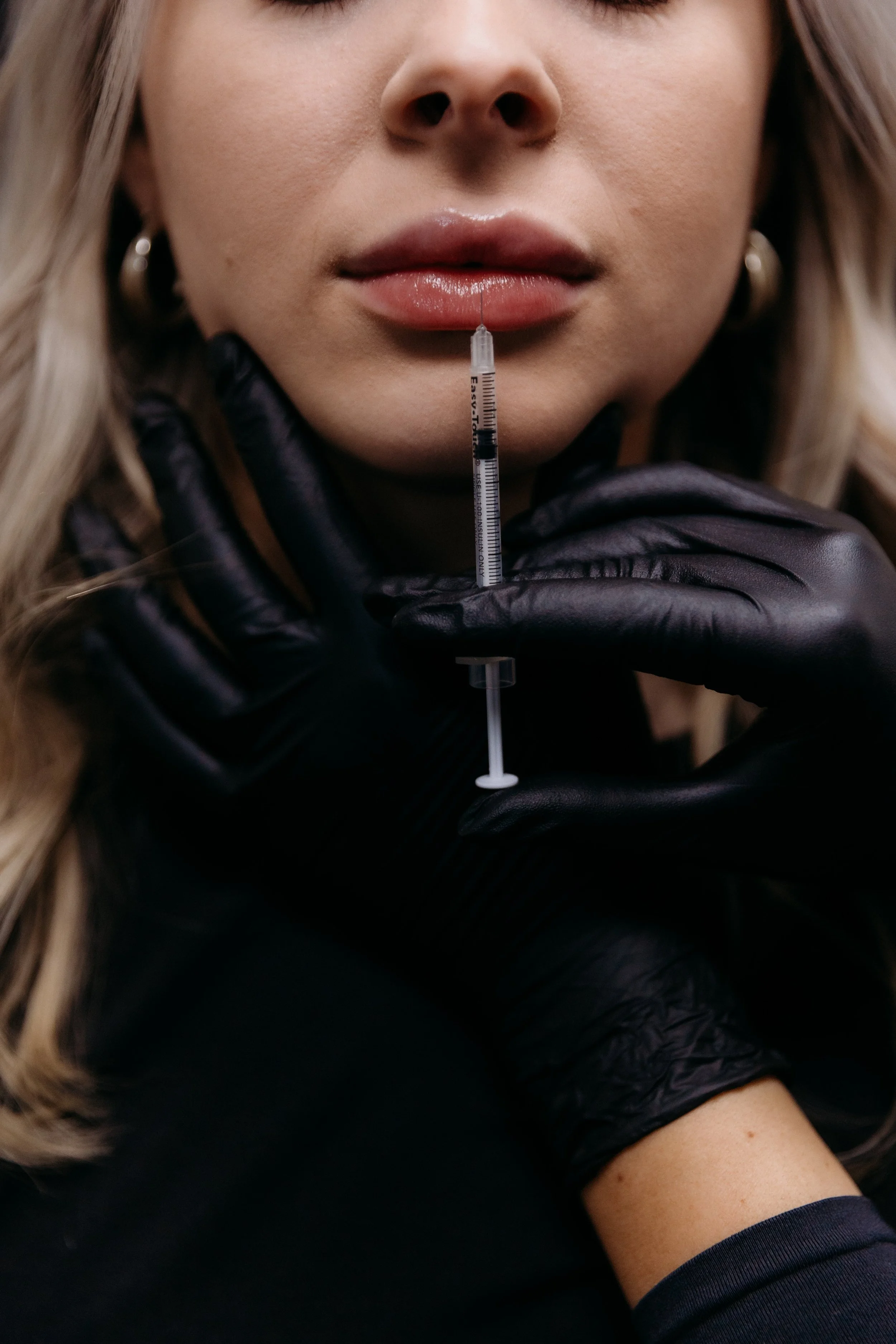 A woman with blond hair is holding a syringe near her lips. She is wearing black gloves and has earrings.
