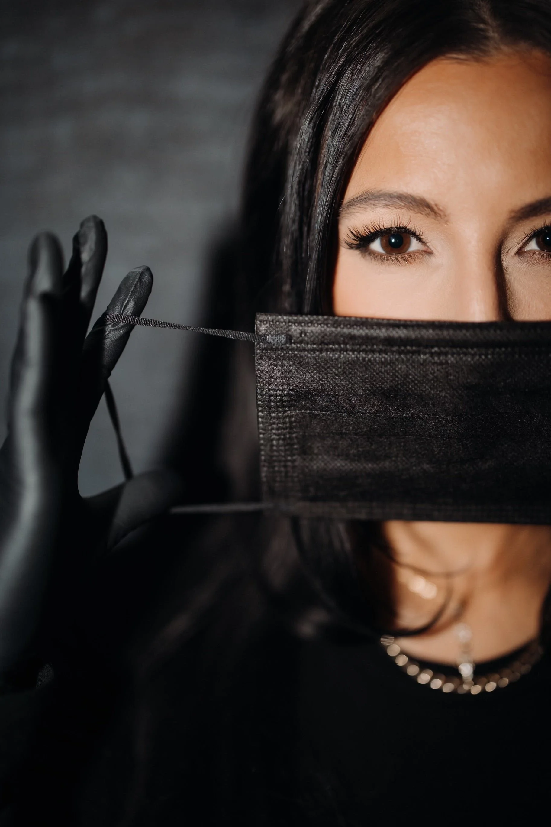 A woman with brown eyes and long dark hair wears a black face mask and black gloves, holding the mask's ear loop with her gloved hand, in front of a dark background.