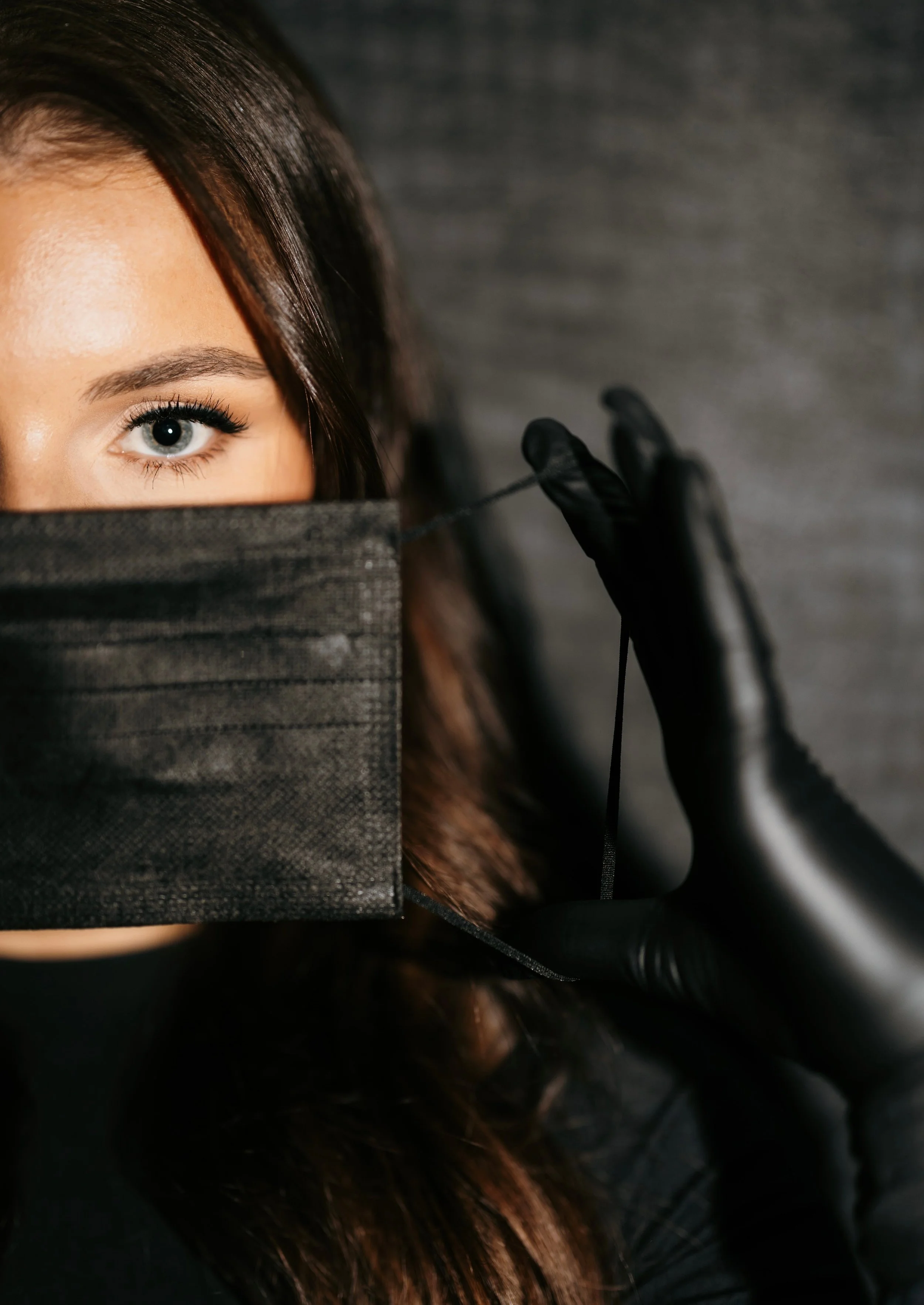 A woman with one eye visible, wearing a black face mask and black gloves, holds the mask's elastic string between her fingers.