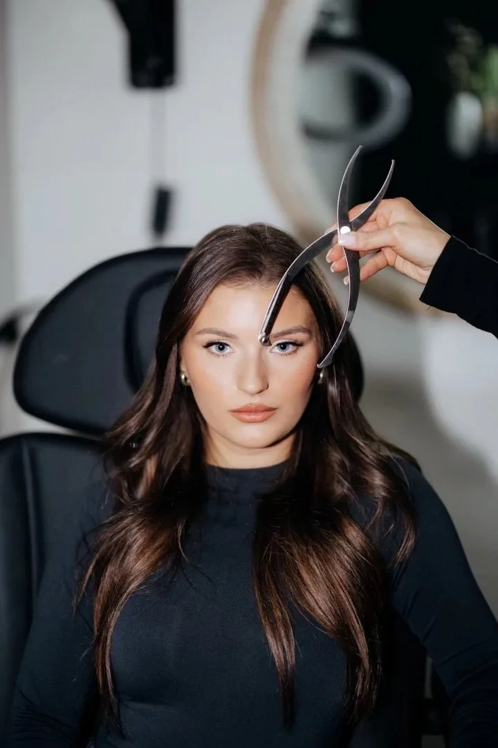 Woman sitting in a chair at a beauty salon with her eyes open as a stylist (not visible) holds eyebrow grooming tools near her face.