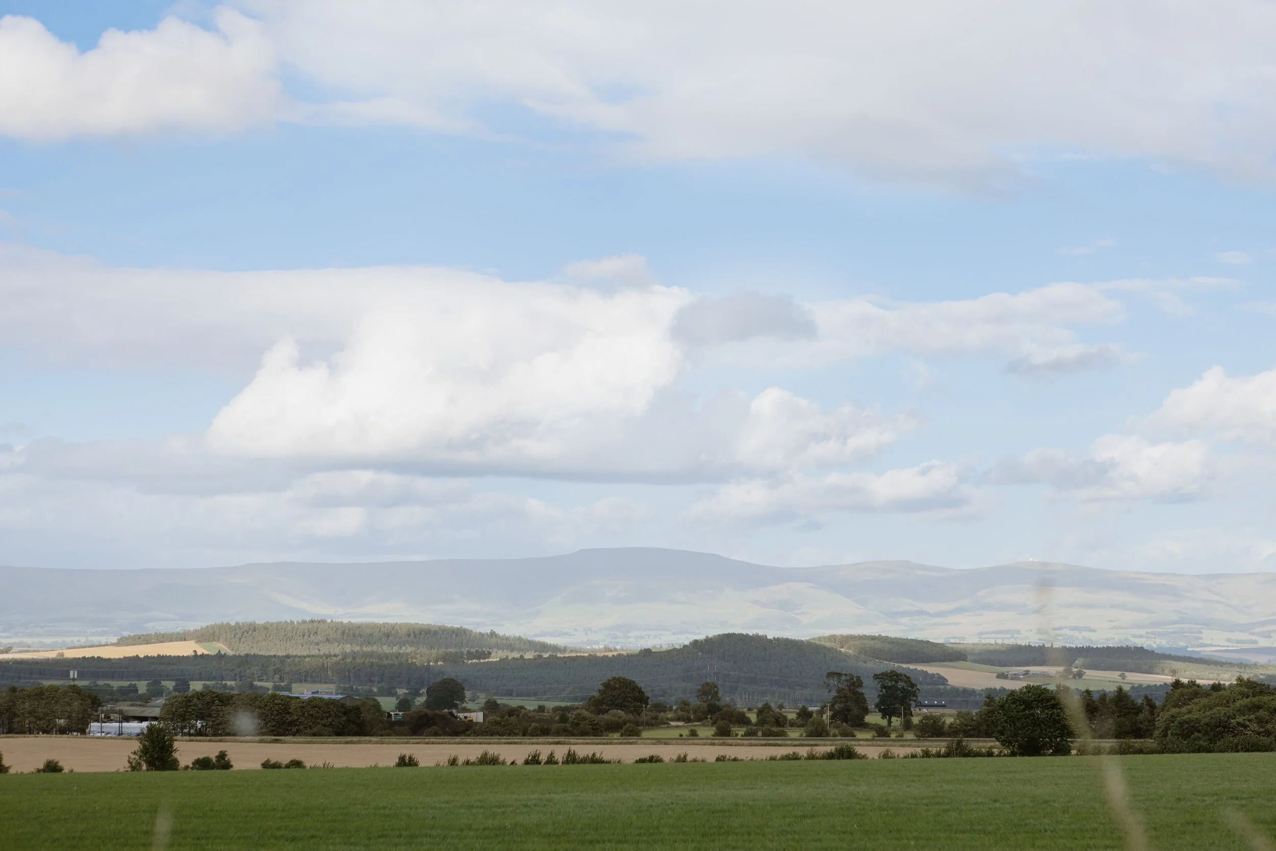 Scenic countryside landscape with open fields, trees, rolling hills, and a partly cloudy sky.