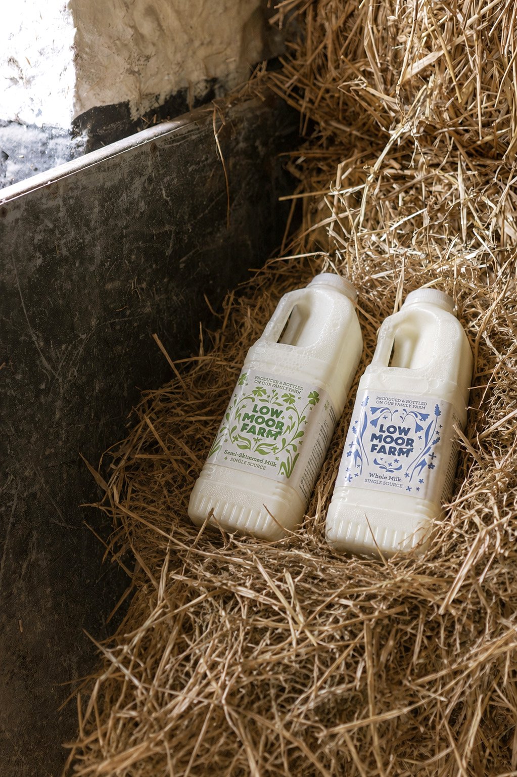 Two bottles of Low Moor Farm milk, one skimmed and one whole, resting on a bed of straw in a barn or farm setting.