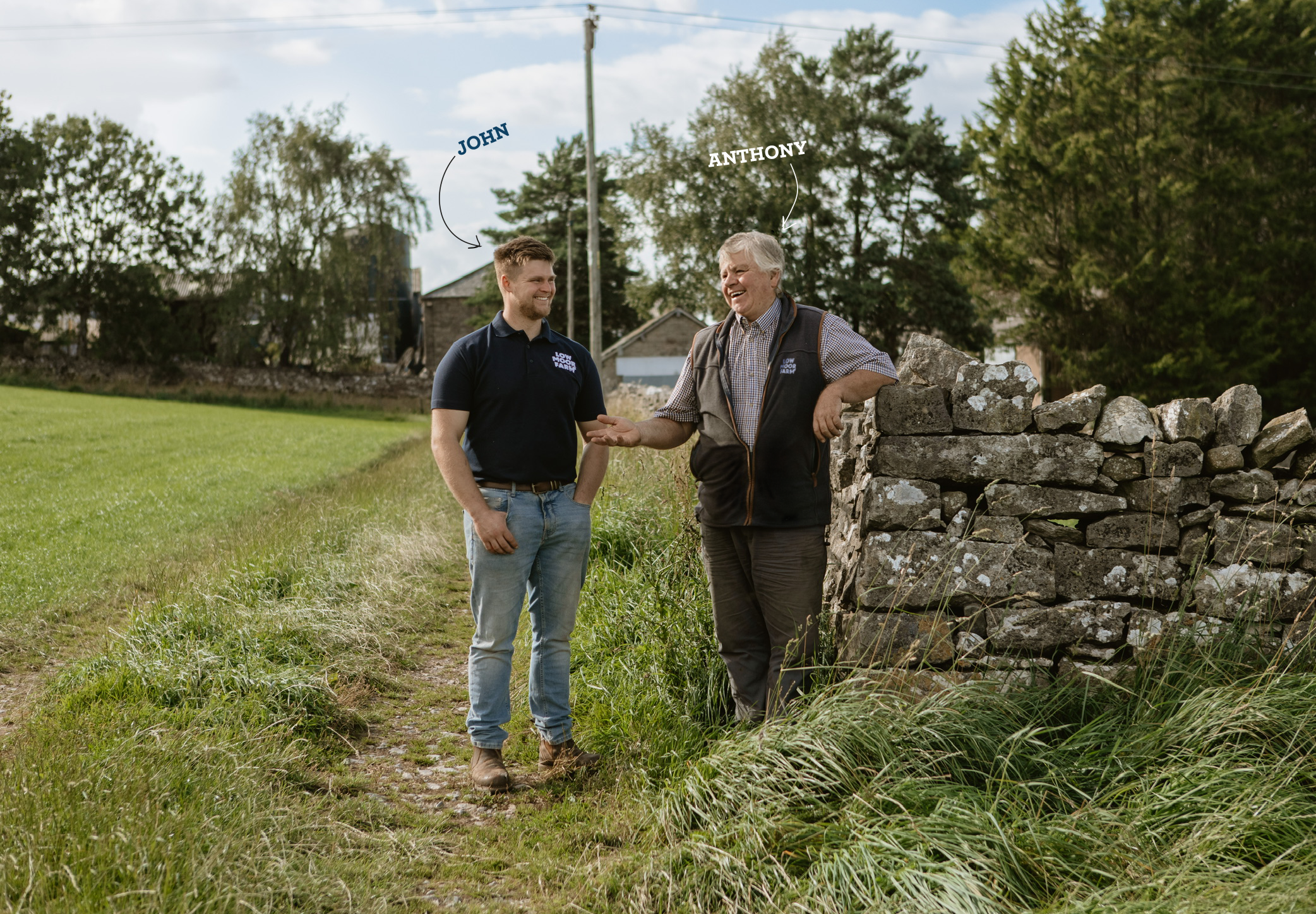 A young man and an older man are standing outdoors near a stone wall, smiling and talking. The young man has short hair and is wearing a black polo shirt and jeans. The older man has gray hair, wears a plaid shirt, a vest, and pants. There is greenery and a few buildings in the background.