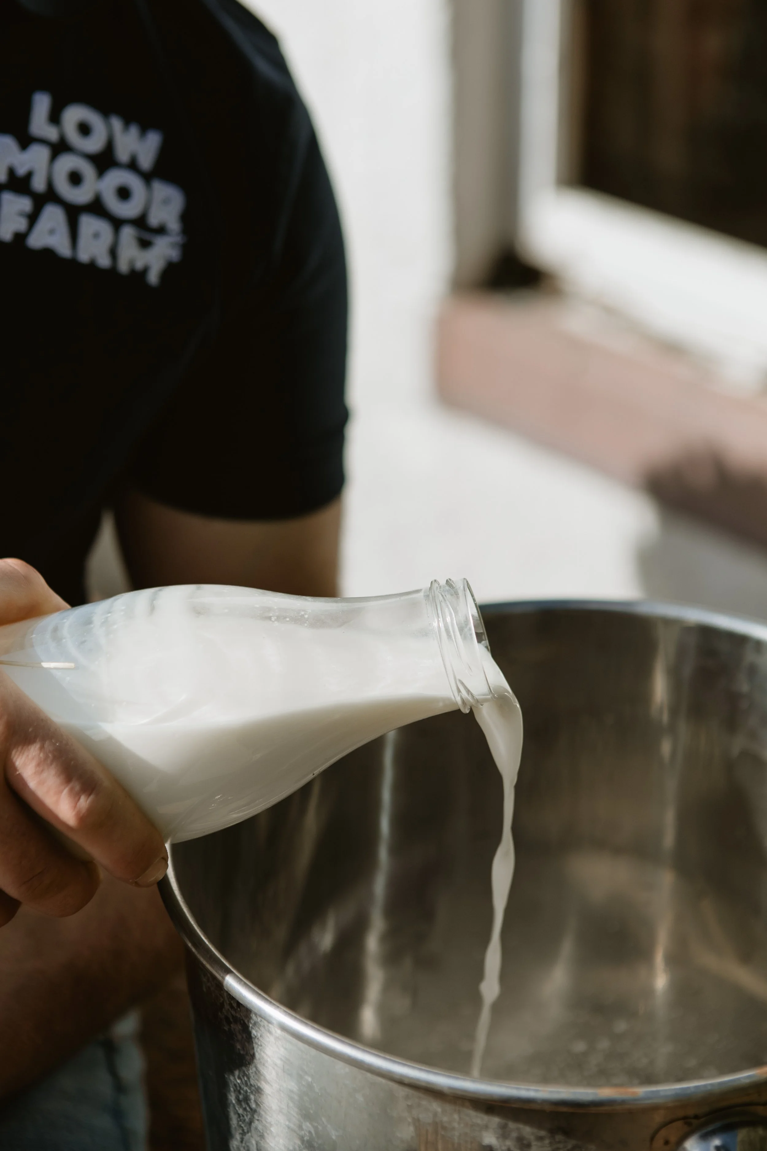Person pouring milk from a glass bottle into a metal container, with a partial view of their black shirt that says 'Low Mood Farm'.