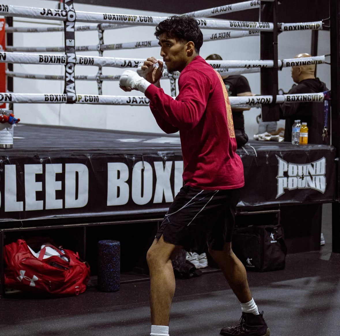 A male boxer in a red shirt and black shorts practicing in a boxing gym. He is standing in a fighting stance near a boxing ring with boxing gloves on, surrounded by equipment and a trainer or staff in the background.