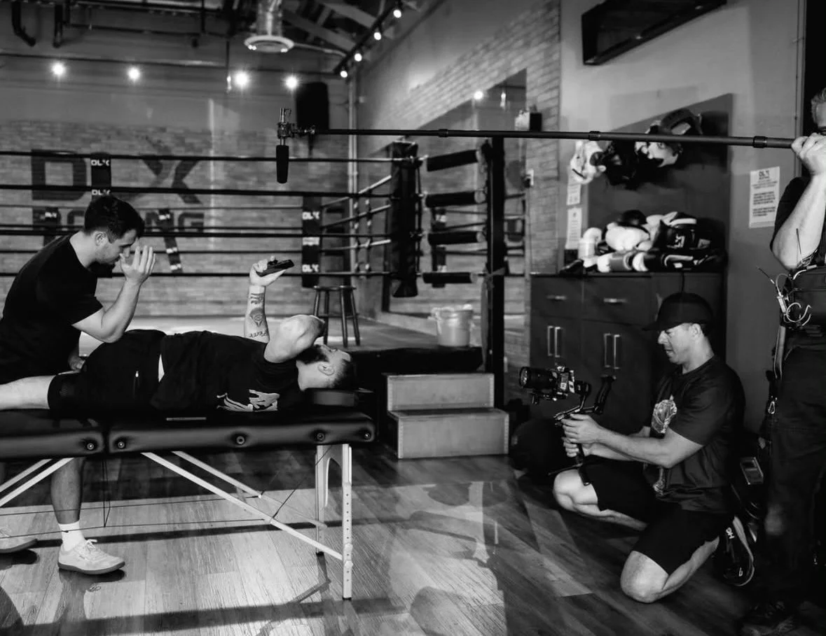 Filming of a recovery session with an athlete lying on a massage table, surrounded by a boom microphone, a camera operator, and a person kneeling on the floor. Boxing gloves and equipment are visible on a cabinet in the background.