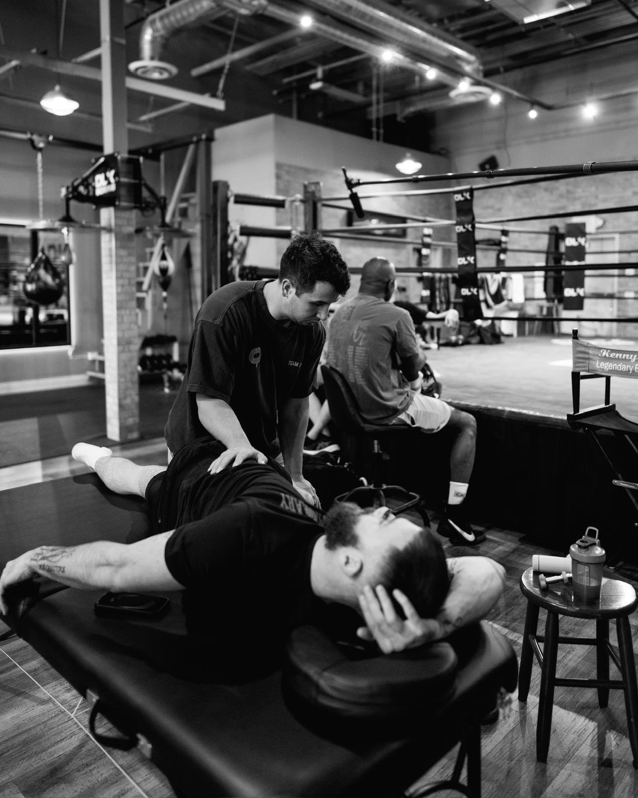A man receiving a chiropractic treatment on his back in a gym, with a boxing ring in the background. Two other people are seated near the ring, one on a stool and the other on a chair. The scene is in black and white.