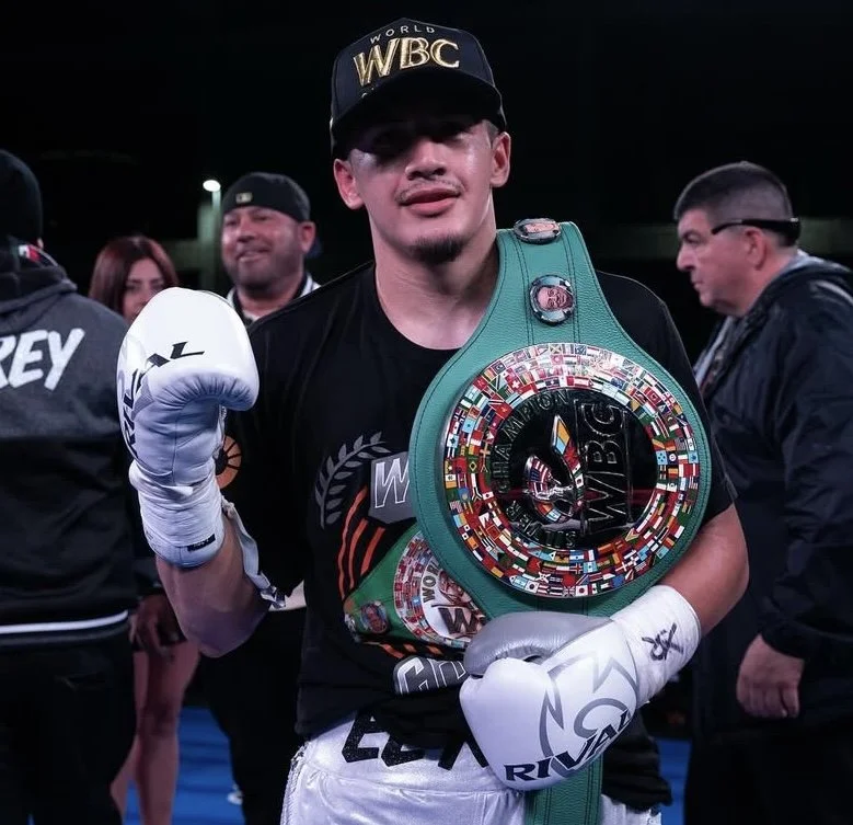 A victorious male boxer wearing a championship belt and cap, posing with clenched fists in a boxing ring surrounded by spectators.