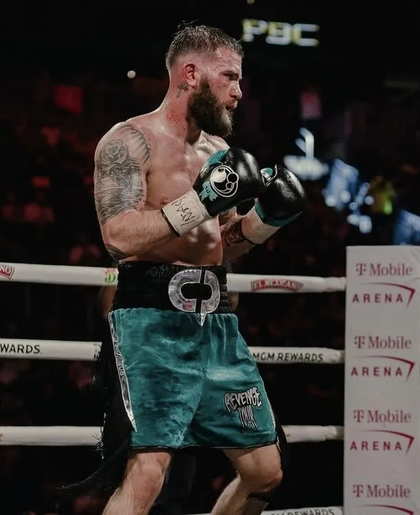 A male boxer with a beard and tattoos on his arms, shirtless, wearing teal shorts and black gloves, standing inside a boxing ring.