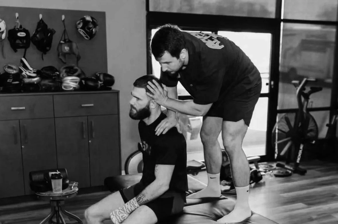 Two men in a gym, one sitting on a mat receiving a head massage from the other standing over him. Gym equipment and accessories are visible in the background.
