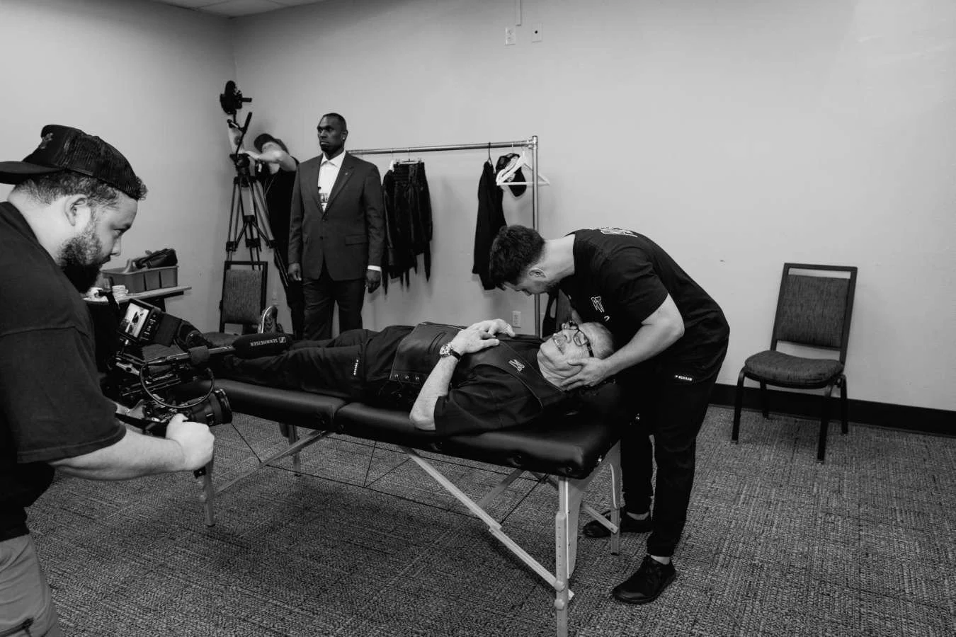 Filming scene with athlete lying on a table, being attended by chiropractor, while another man stands in the background with a camera and director's headset.