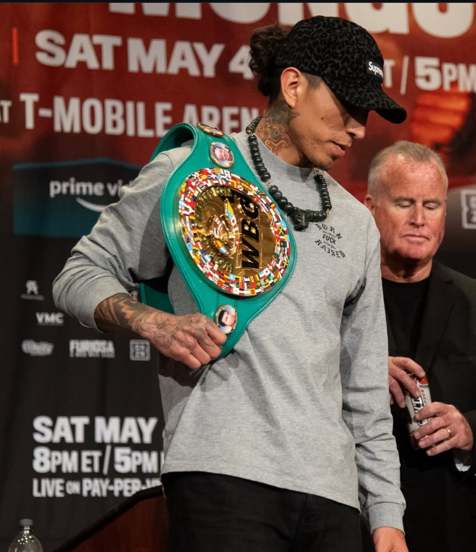 A man with tattoos and a cap, wearing a gray sweatshirt, holds a colorful WBC boxing championship belt over his shoulder. An older man stands beside him, holding a microphone or spray can, in front of a promotional backdrop for a fight event scheduled for Saturday, May 4.