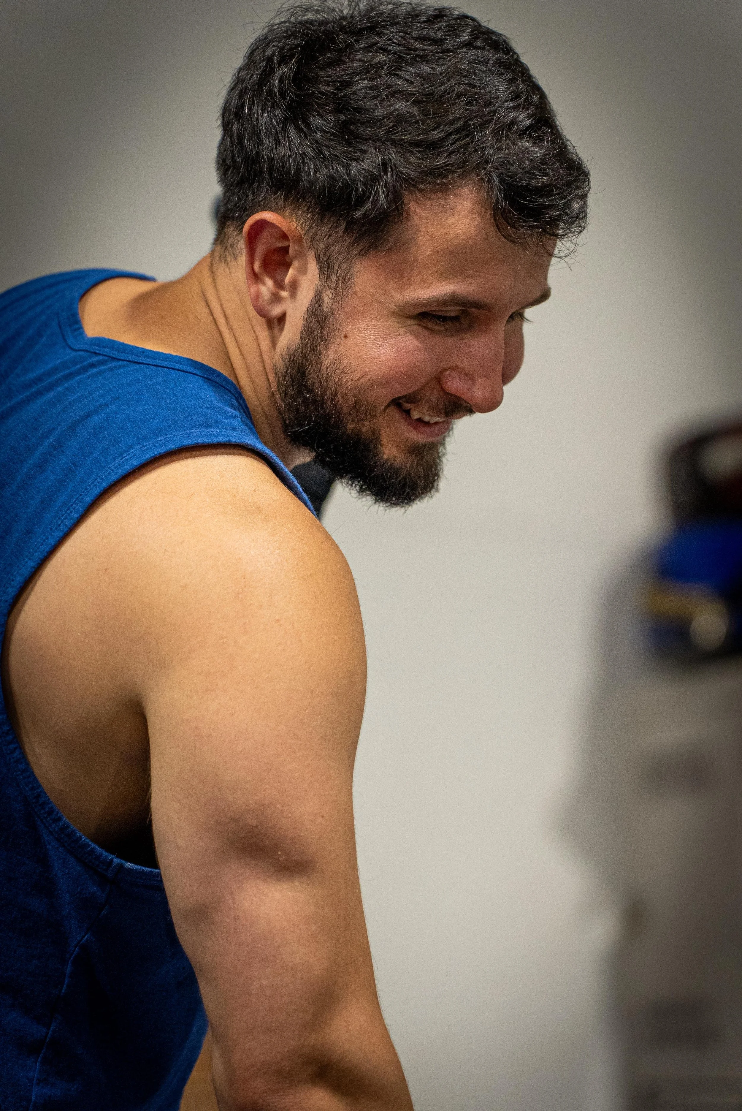 A young man with dark hair and a beard, smiling, wearing a blue sleeveless athletic shirt, indoors against a plain wall.