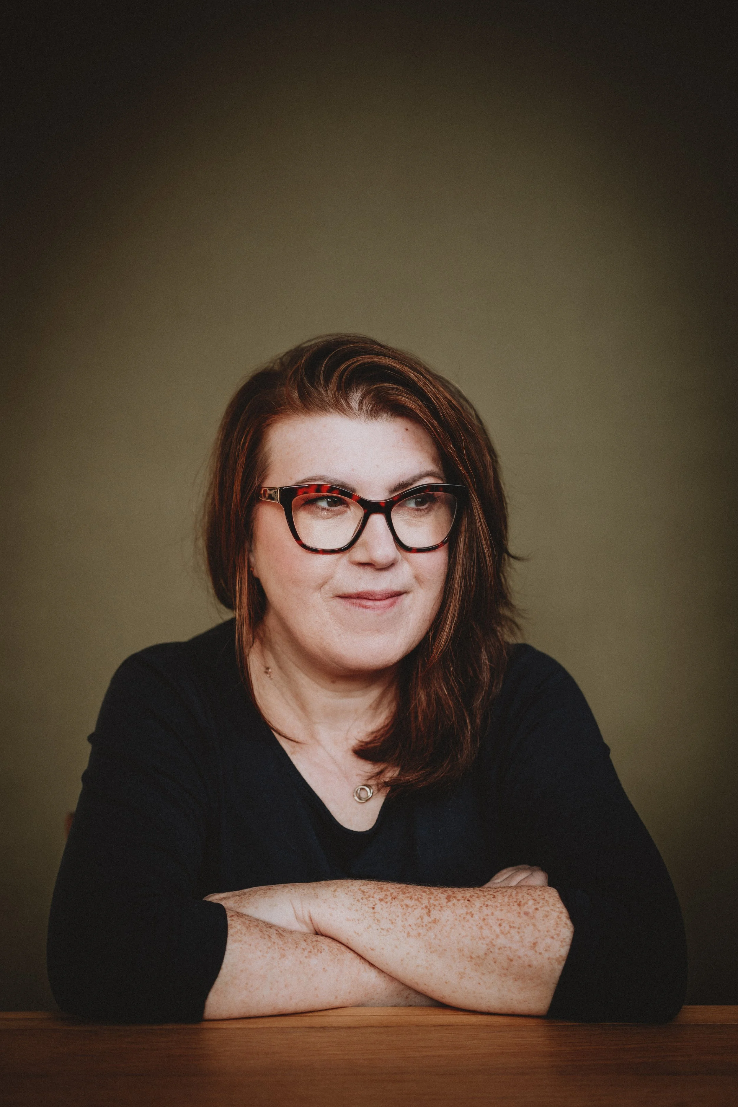 A woman with red hair, glasses, and freckles sitting at a wooden table with her arms crossed, smiling softly, against a plain beige background.