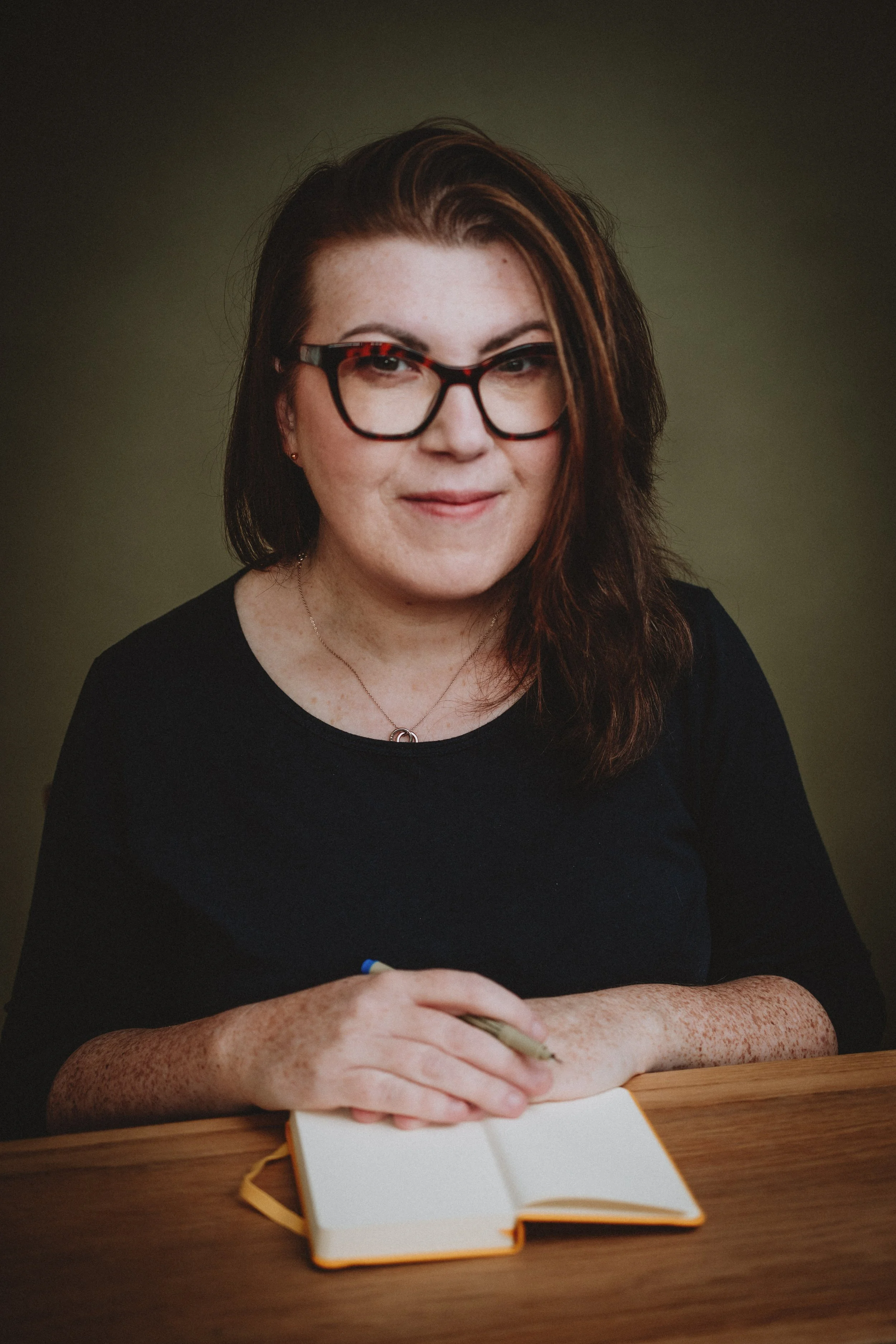A woman with glasses, brown hair, and freckles sitting at a wooden table with an open notebook in front of her, holding a pen.