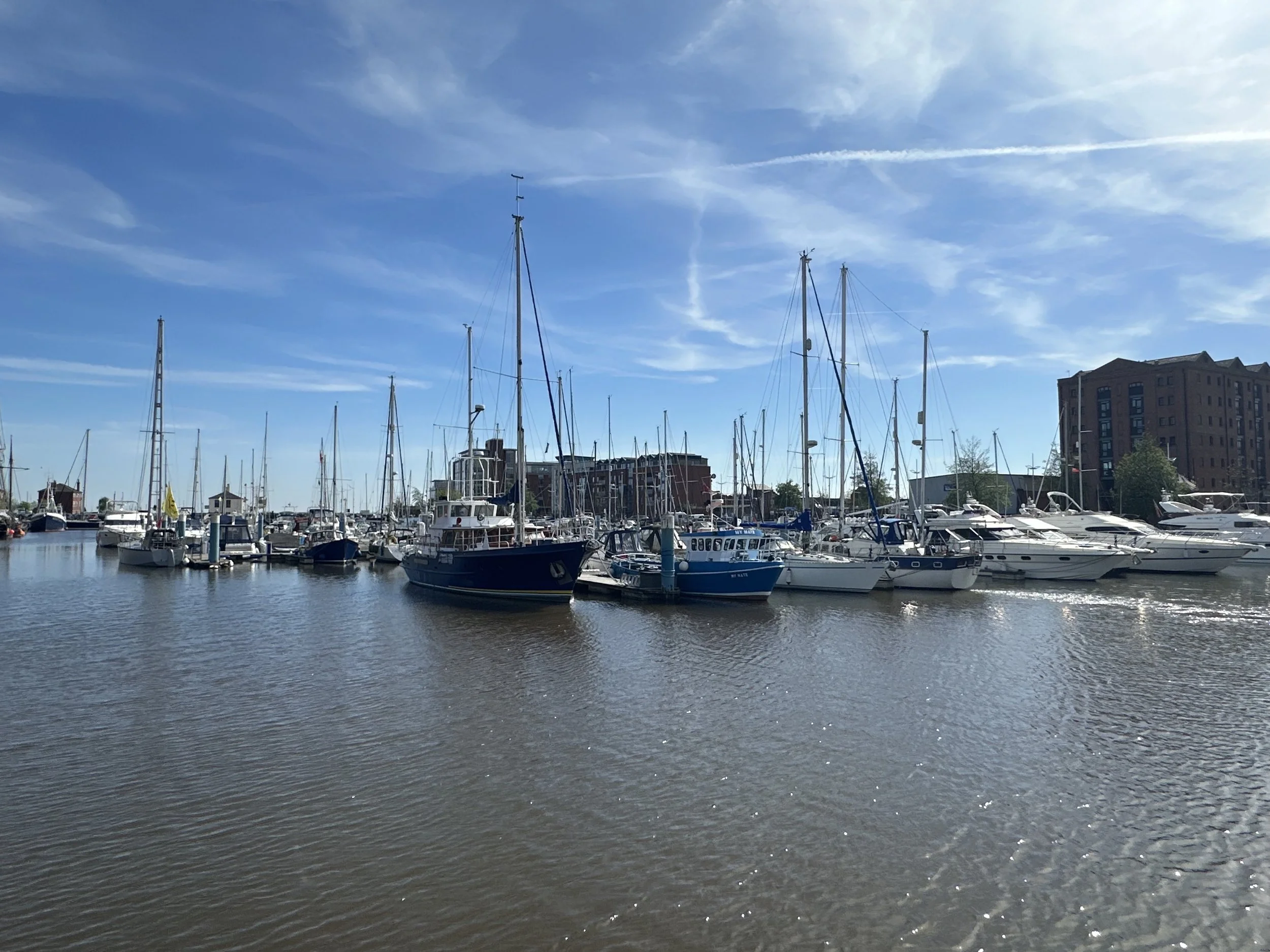 Marina with various sailboats and yachts docked on the water, multi-story buildings in the background, partly cloudy sky with contrails.