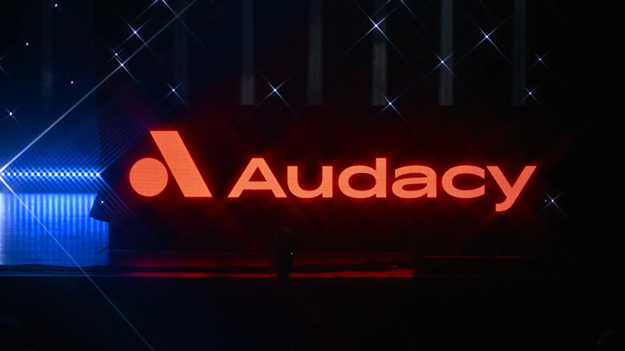 Bright red illuminated Audacy logo and text on a dark stage with spotlights and star-like light reflections.