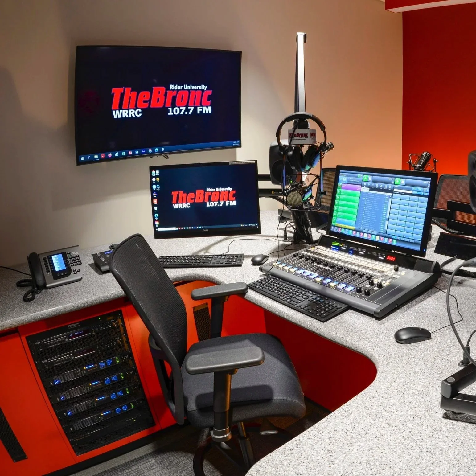 Radio broadcasting studio with desk, computer monitors displaying the logo for The Bronc, headphones, audio equipment, and a chair.