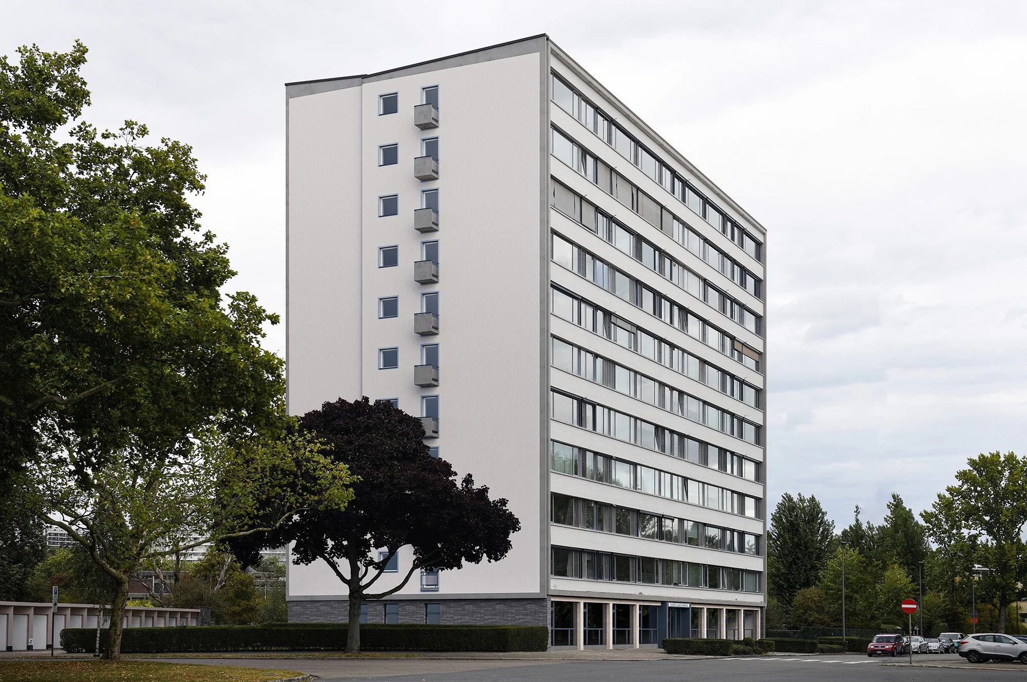 A tall, white multi-story residential building with many windows and small balconies. The building is surrounded by trees and parked cars, with a cloudy sky overhead.