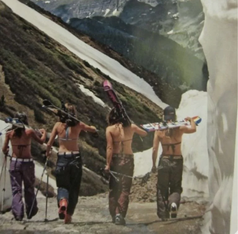 Four women with ski equipment walking through a mountain landscape with snow and glacier, carrying skis on their shoulders.