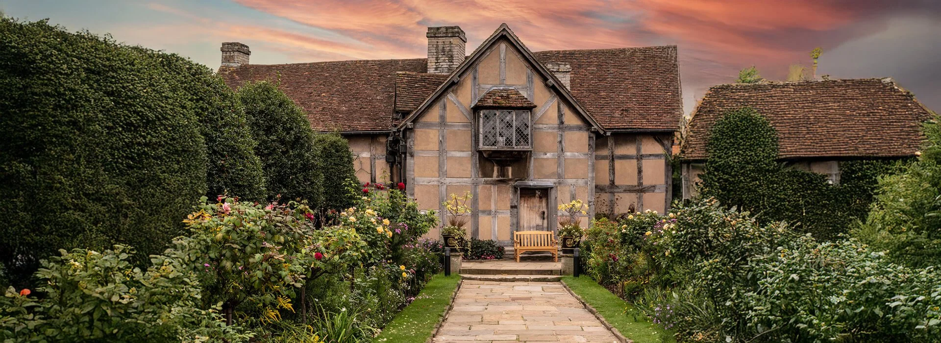 Lush English cottage garden at sunset, with vibrant resilient perennial and rose borders, clipped hedges, and a stone path leading to a bench in front of a historic timber-framed house.