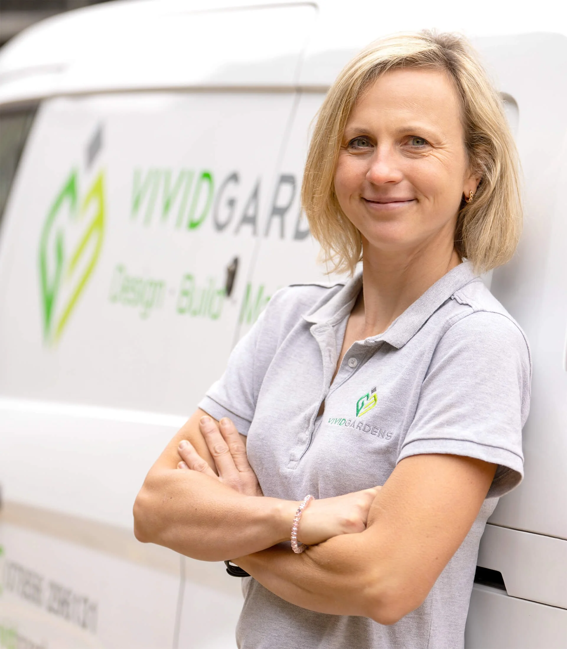 Woman in a Vivid Gardens polo shirt standing with arms crossed in front of a Vivid Gardens van, promoting landscaping services in Buckinghamshire, Hertfordshire, and Oxfordshire.