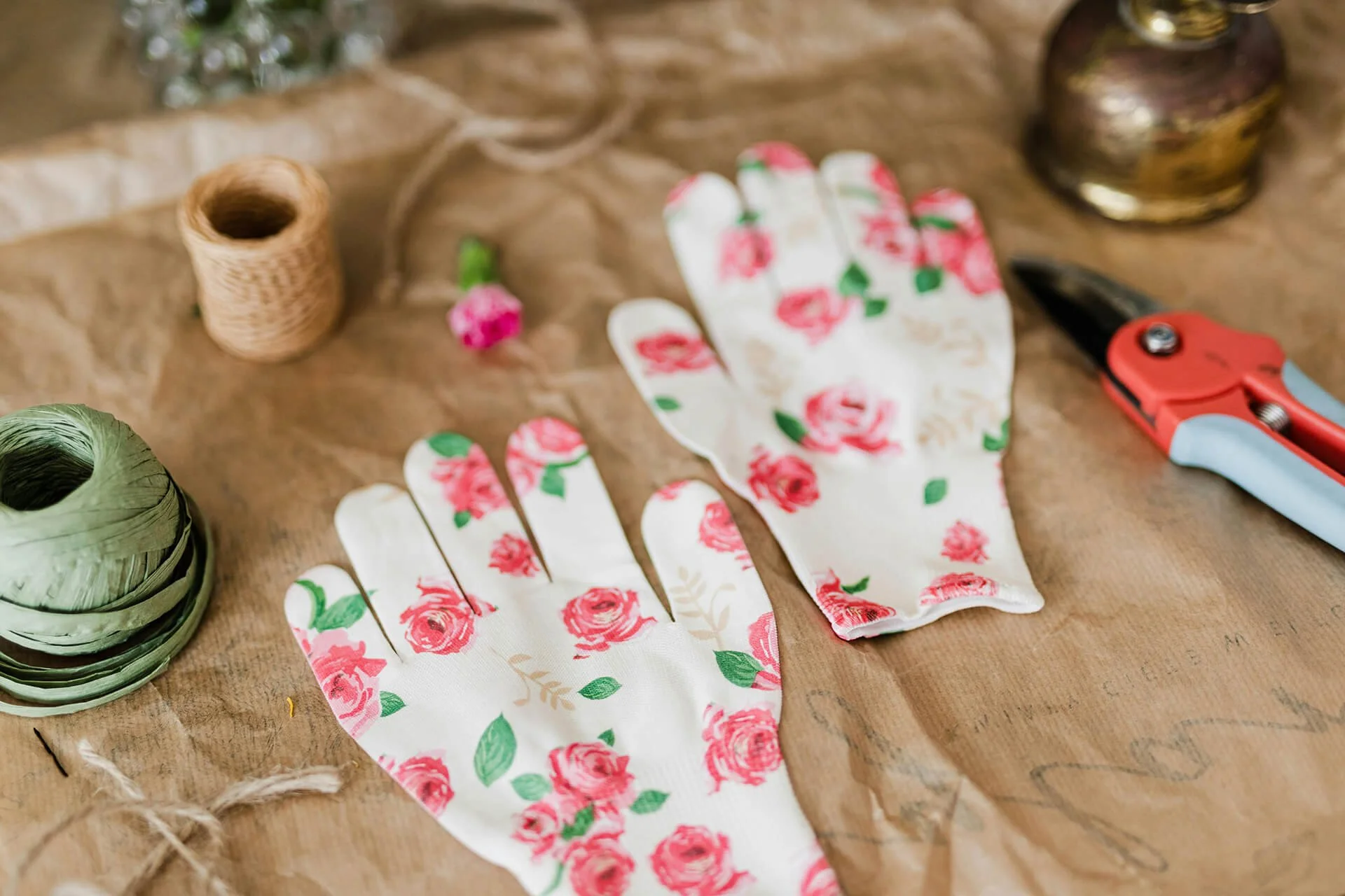 A pair of gardening gloves with a floral pattern, a pair of scissors, spools of twine, and a small flower on a wooden surface for crafting or gardening.
