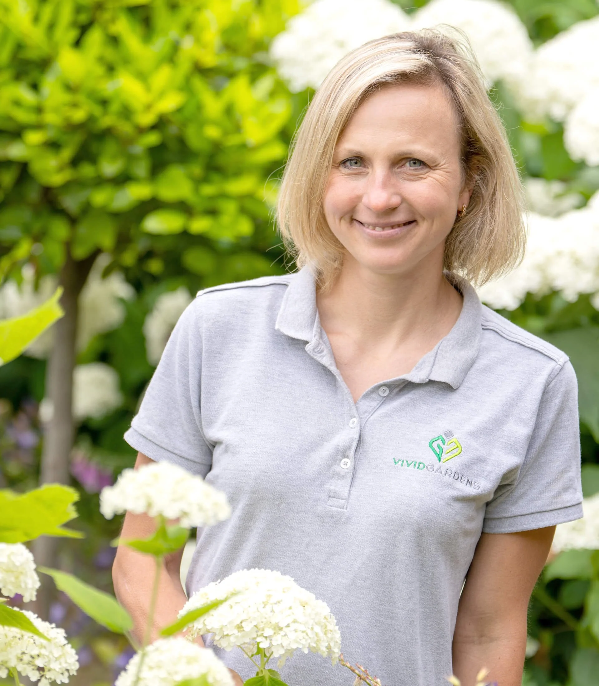 A smiling woman in a gray Polo shirt with 'Vivid Gardens' logo, standing outdoors among white flowering plants and green foliage.