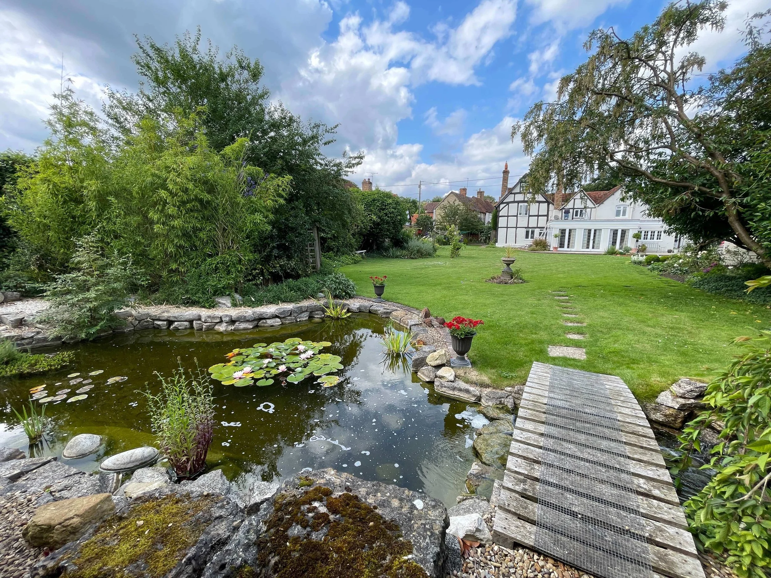 A backyard with a pond filled with water lilies, surrounded by rocks and lush greenery, with a small wooden bridge crossing over the pond, a manicured lawn, flower pots, stepping stones, and houses with white and black exterior in the background under a partly cloudy sky.