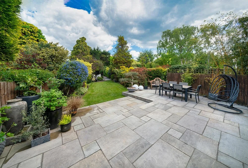 Spacious stone patio with outdoor dining set overlooking lush landscaped garden with colorful shrubs and manicured lawn
