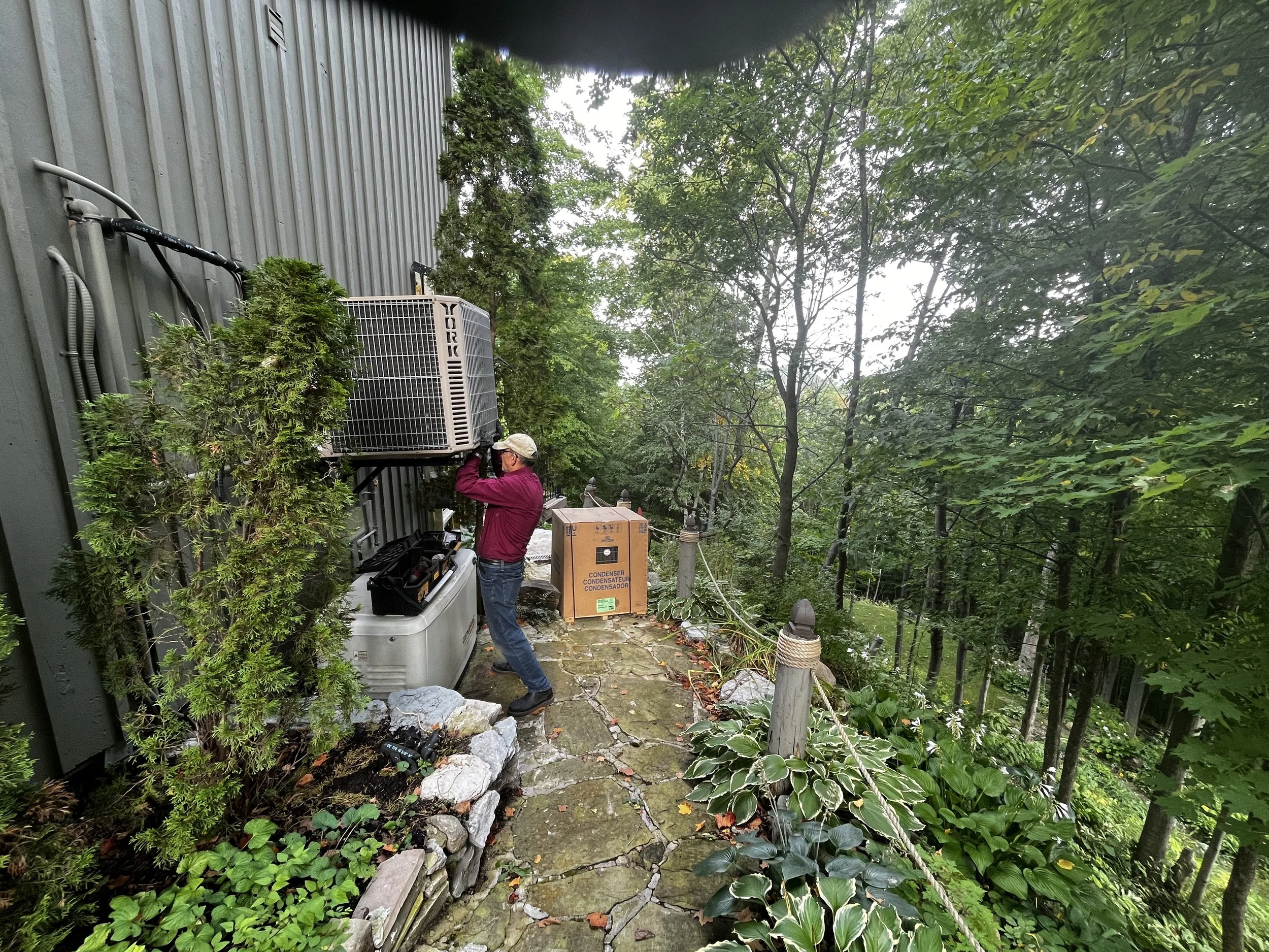 A man in a burgundy jacket and beige cap works on an outdoor air conditioning unit mounted on the side of a building, surrounded by green trees and plants.