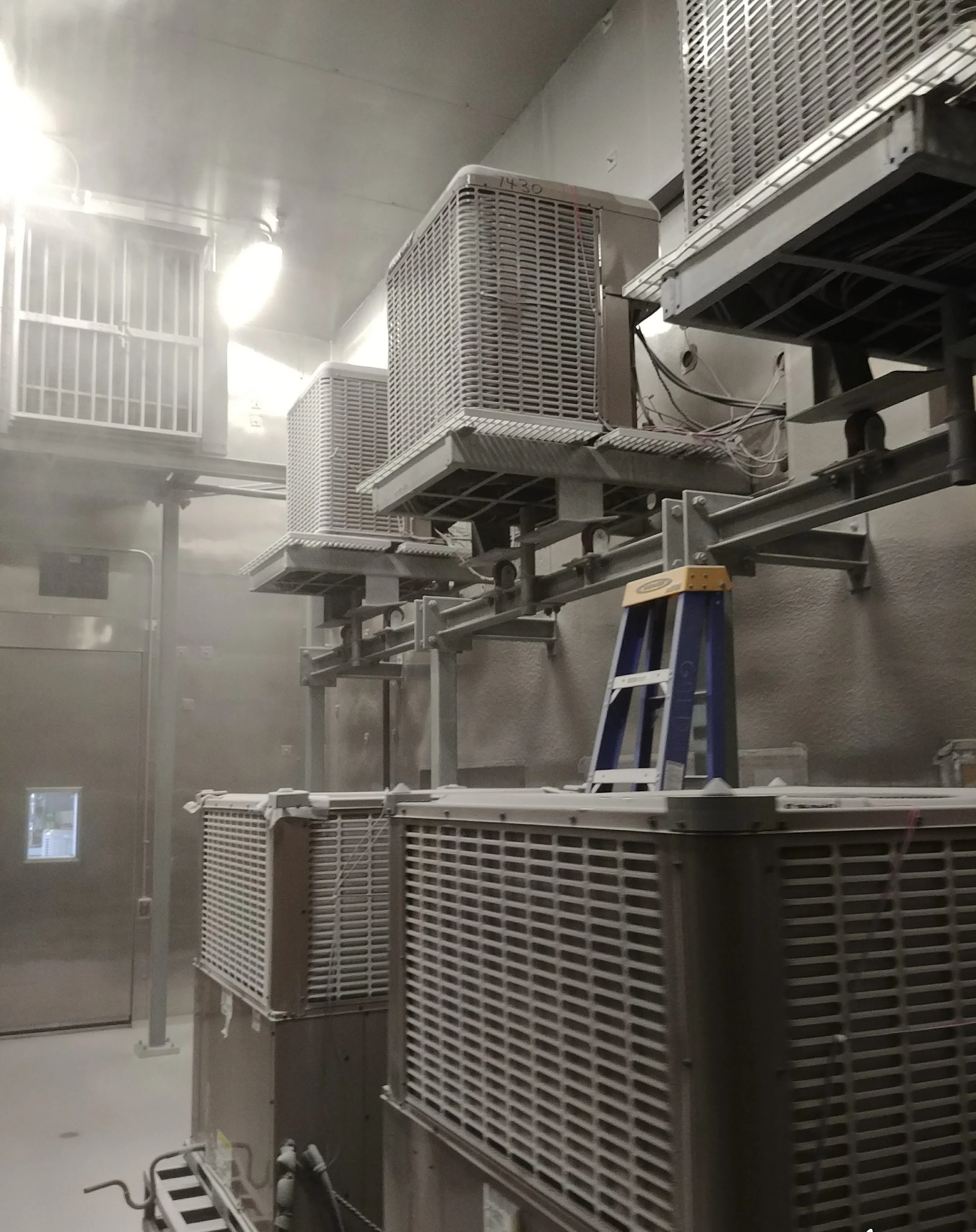 Industrial HVAC units installed on a beige wall, with a blue step ladder nearby and a mist or fog present in the room.