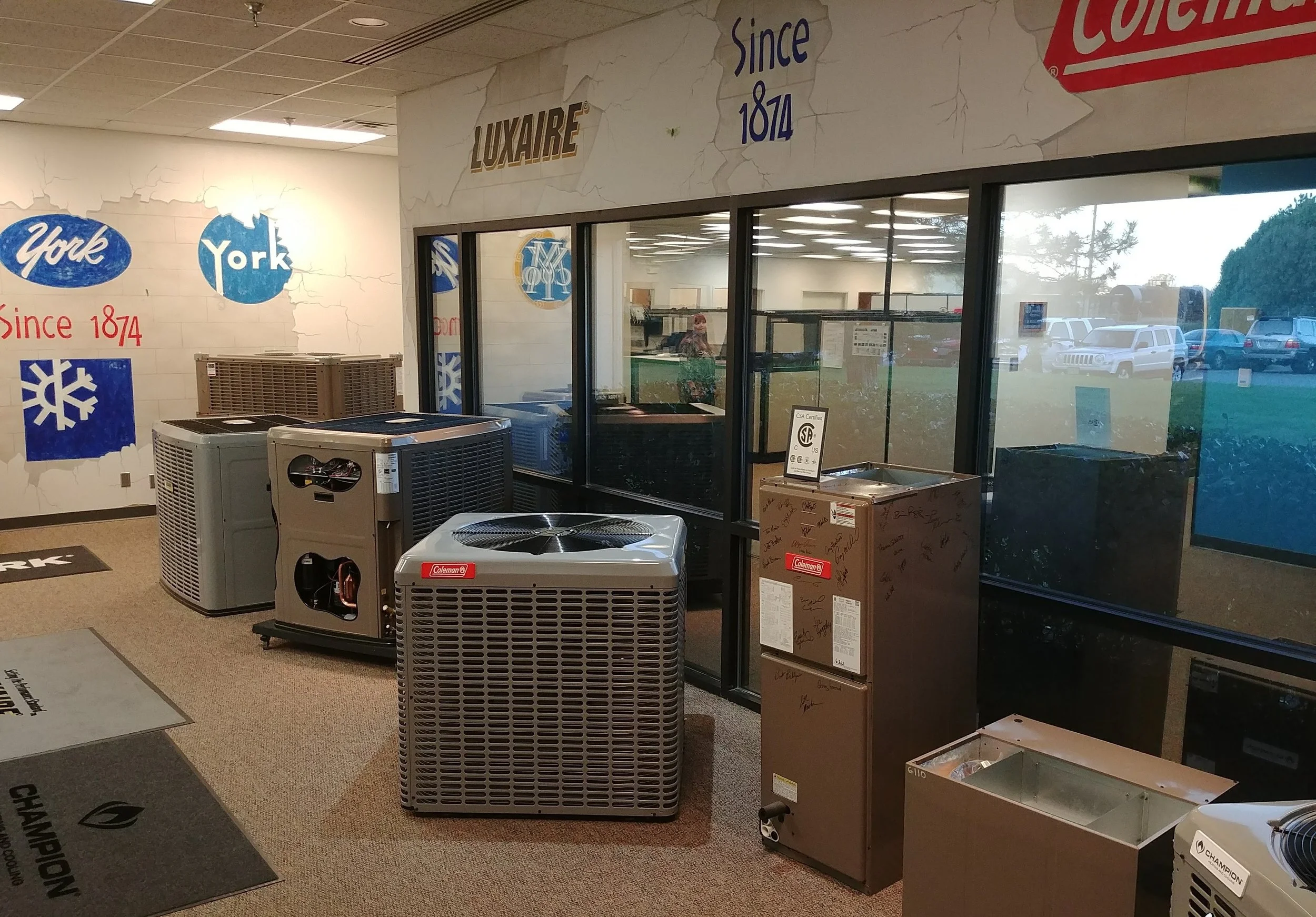 Indoor display area with multiple HVAC units, signage indicating the store since 1874, and large glass windows showing parked cars outside.