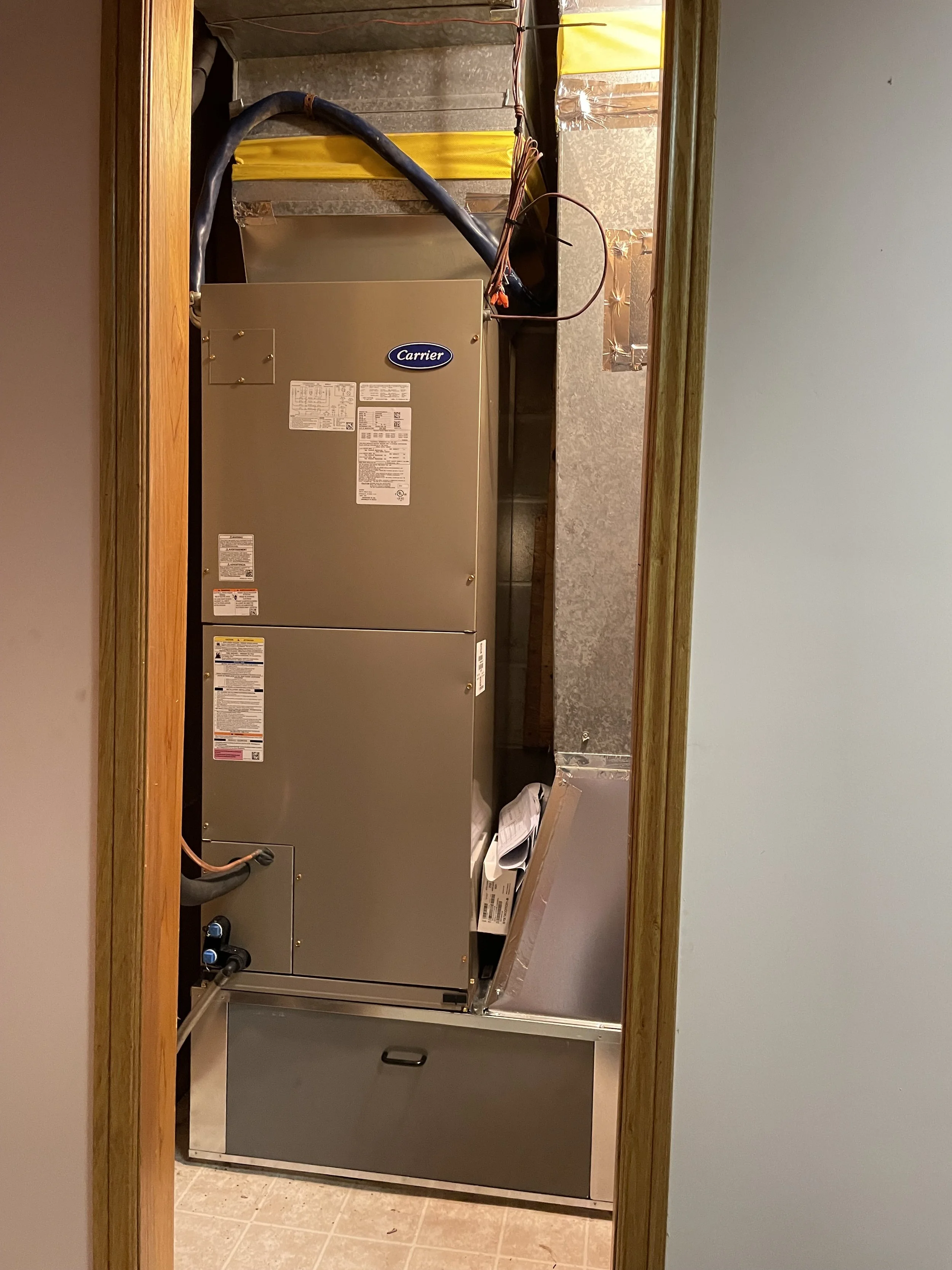 View of a beige Carrier HVAC unit installed in a small closet with unfinished walls, surrounded by pipes and wires.