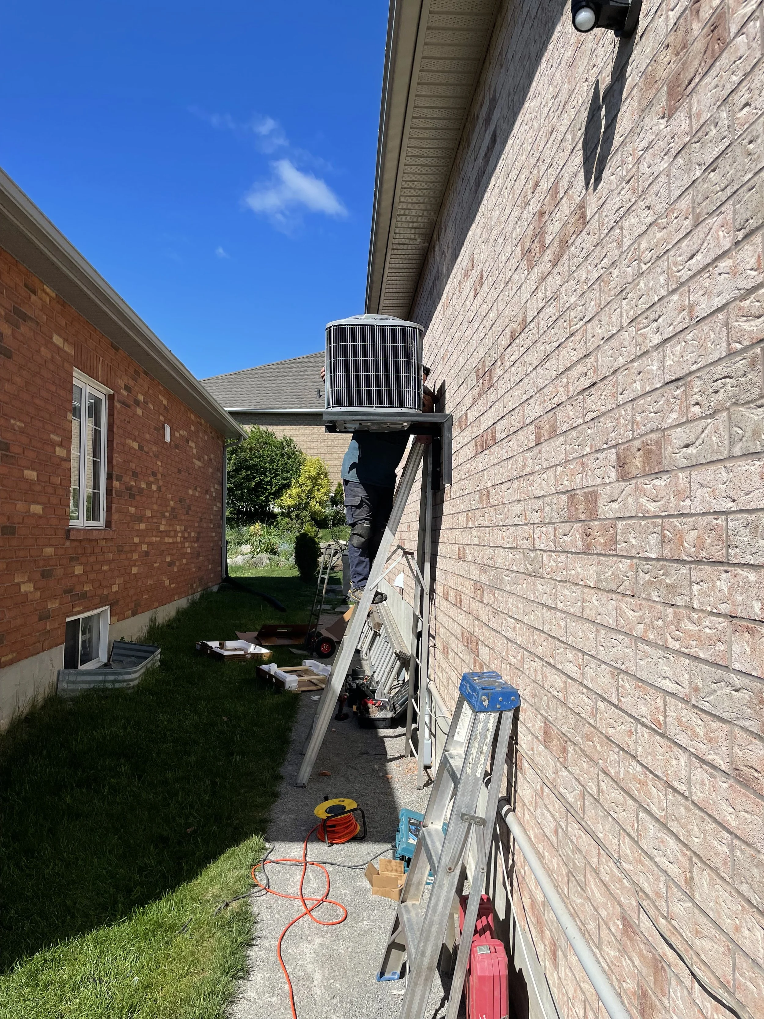A worker on a ladder repairing or installing an air conditioning unit outside a brick house on a sunny day.