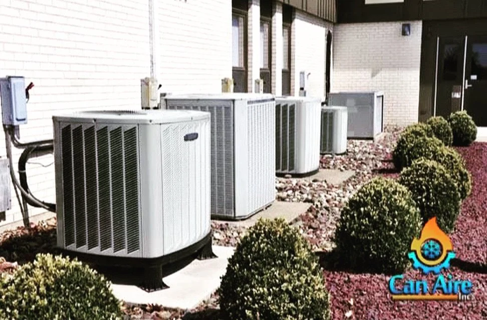 Multiple air conditioning units are installed outside a building, surrounded by small green bushes and decorative rocks.