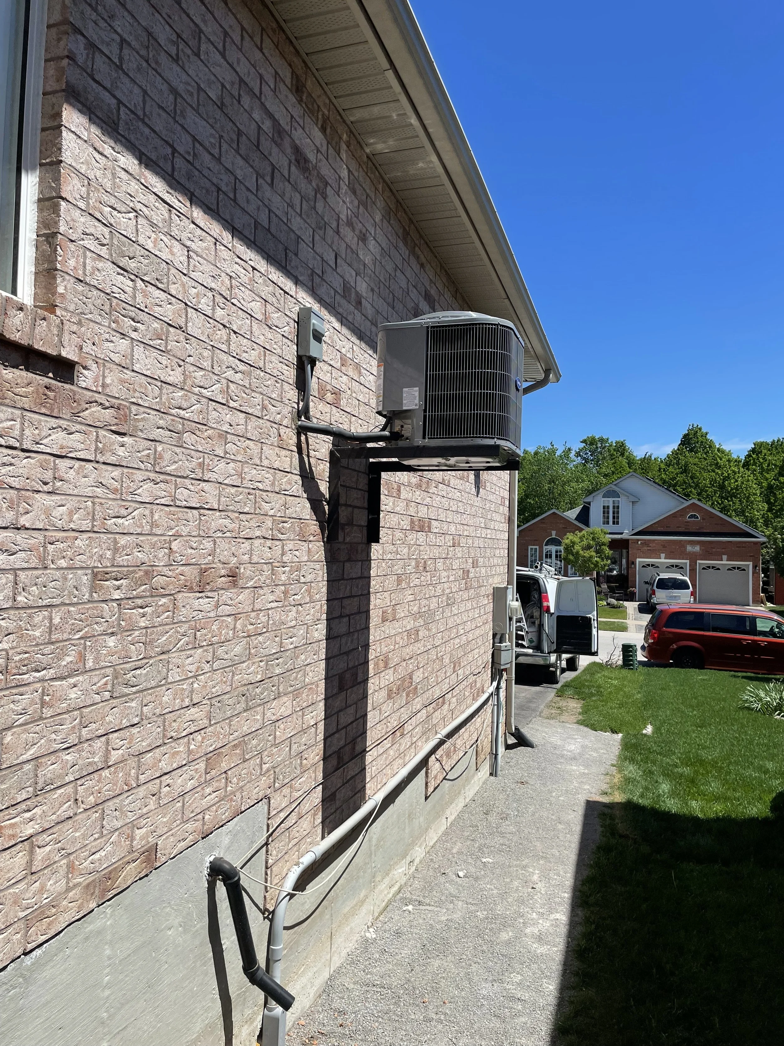 Exterior view of a brick house with centralized air conditioning unit mounted on the wall and connected to electrical and plumbing systems, in a suburban neighborhood with parked cars and green lawns under a clear blue sky.