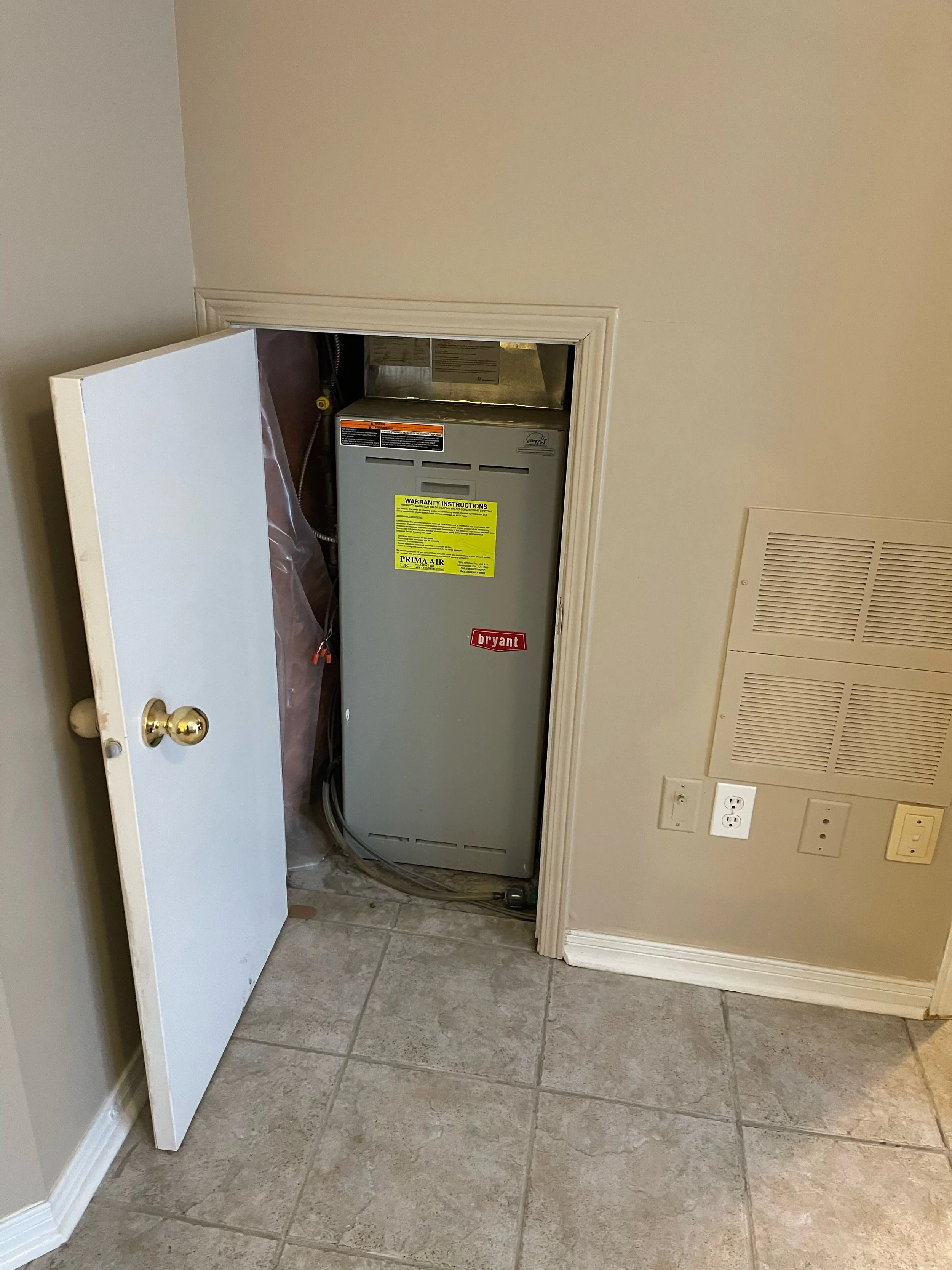 A small utility closet with a Bryant furnace installed inside, partially concealed by a white door that is open, with beige tiled flooring and a beige wall with electrical outlets and a vent.