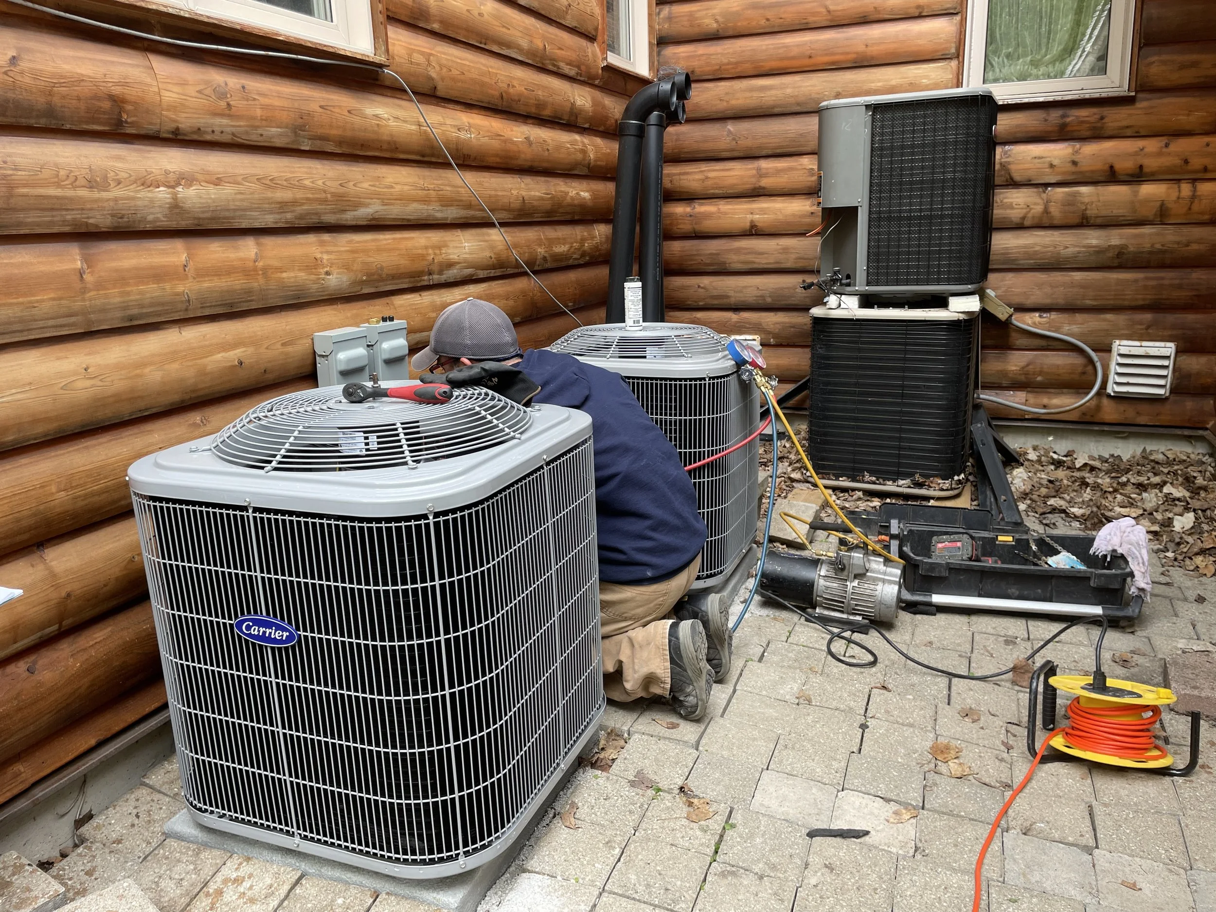 A person kneeling outside next to two air conditioning units, working on maintenance or repair, with tools like a wrench and a toolbox nearby, on a stone patio beside a wooden house wall.