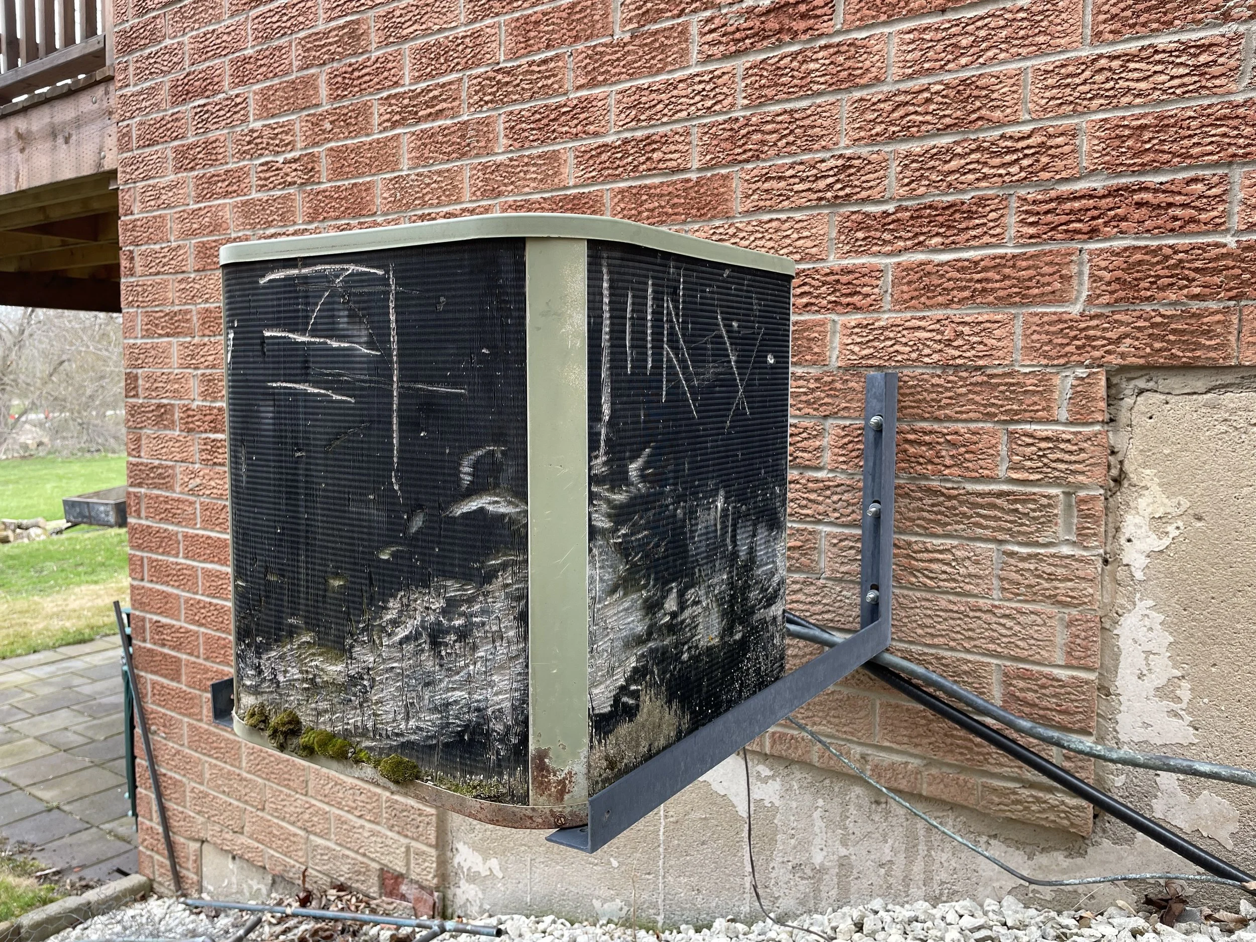 The outdoor unit of an air conditioning system mounted on an exterior brick wall, with visible signs of mold, dirt, and worn paint.