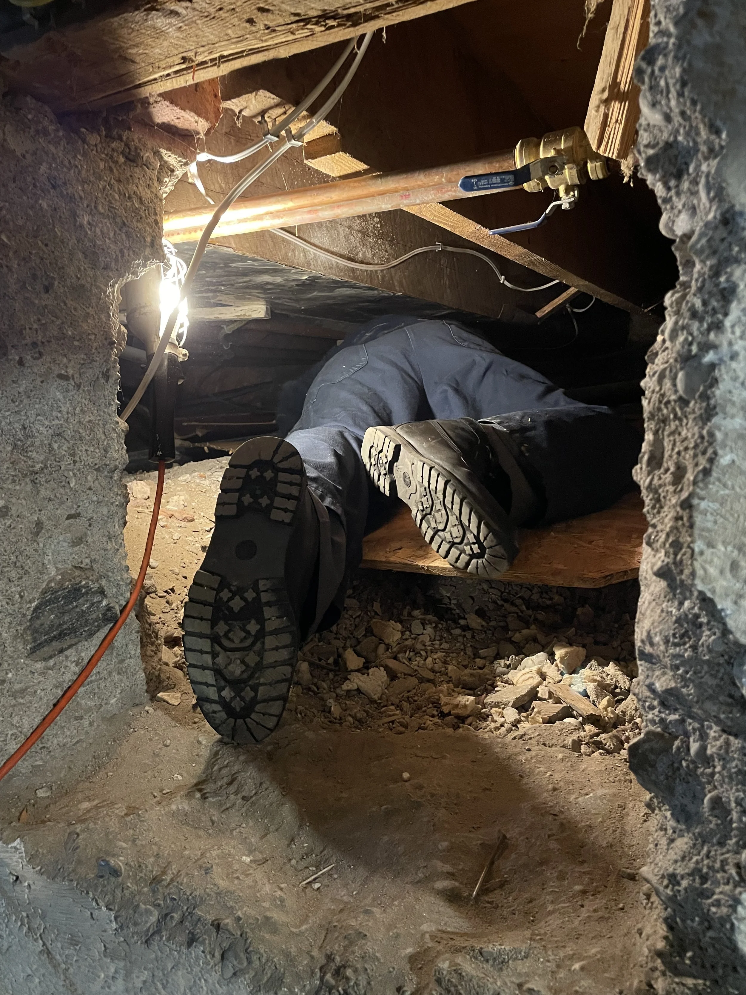 A person crawling through a confined space or crawl space underneath a building or house, with exposed wiring and pipes visible above.